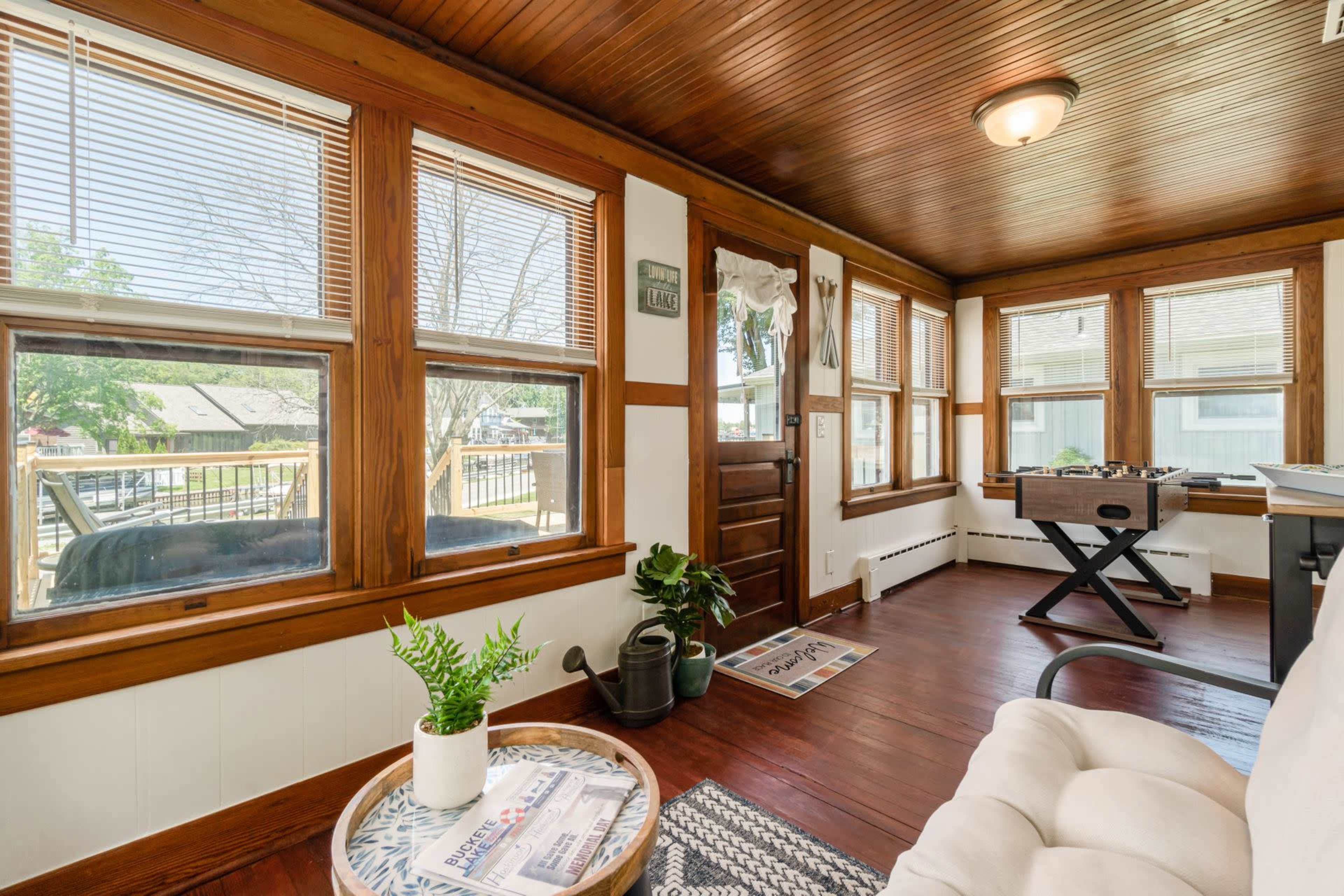 A sunlit room with wooden ceiling and trim, featuring large windows, a comfortable chair, and a small table with a plant.