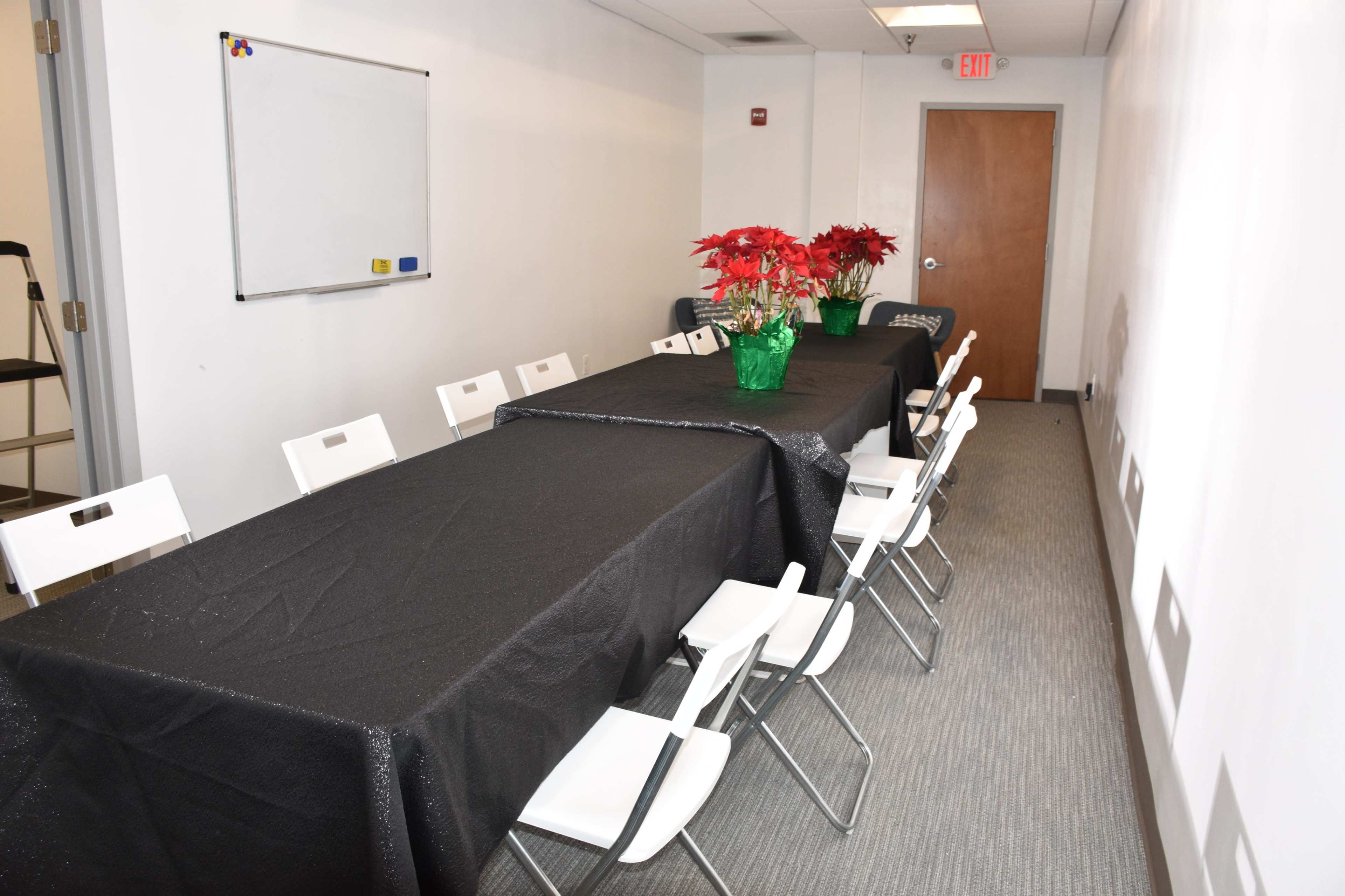 A long table covered with a black tablecloth is set in a conference room, lined with white chairs, and adorned with a centerpiece of potted red poinsettias.