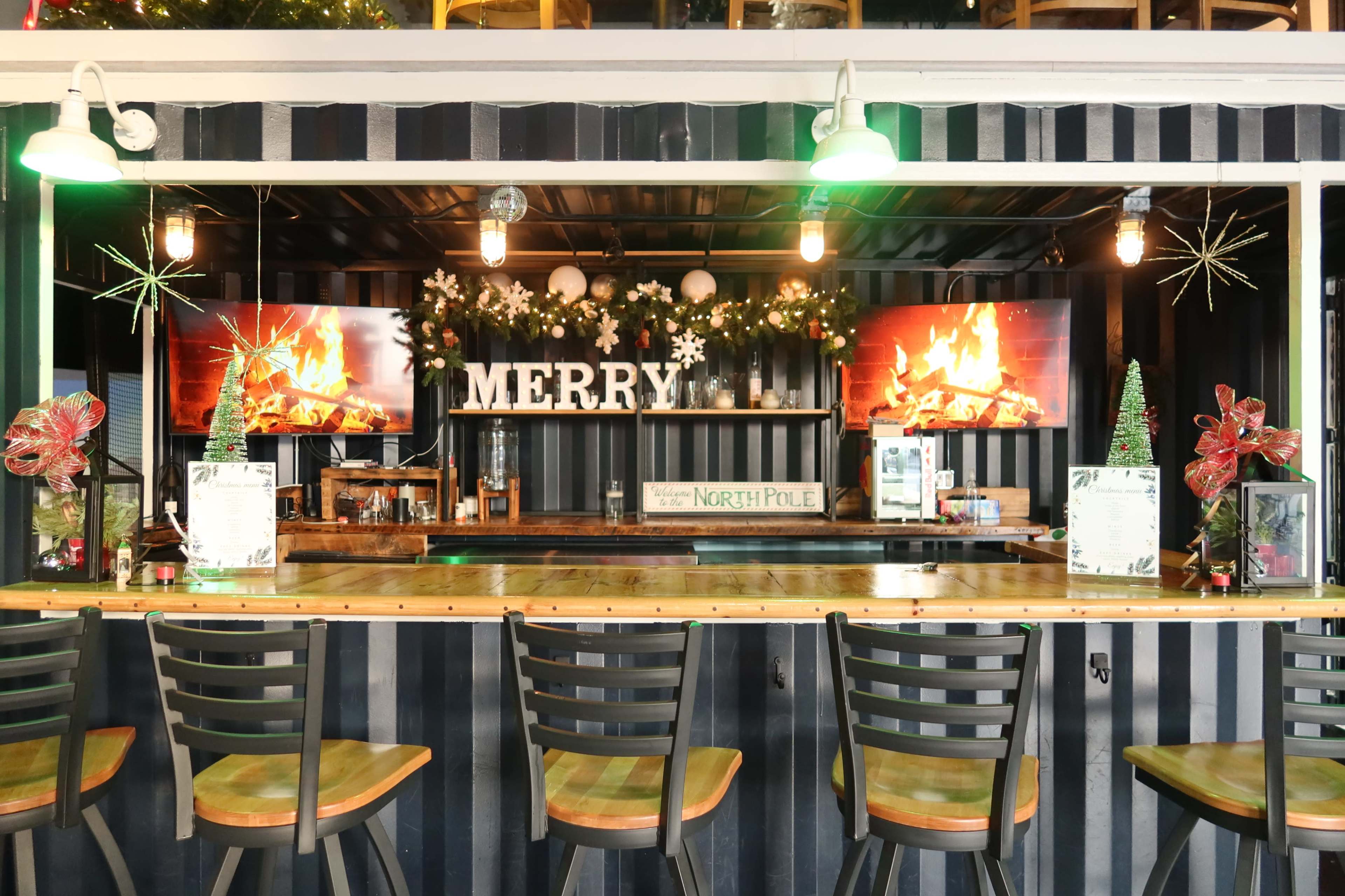 The image shows a bar area decorated with a "MERRY" sign, holiday garlands, and two screens displaying a virtual fireplace.