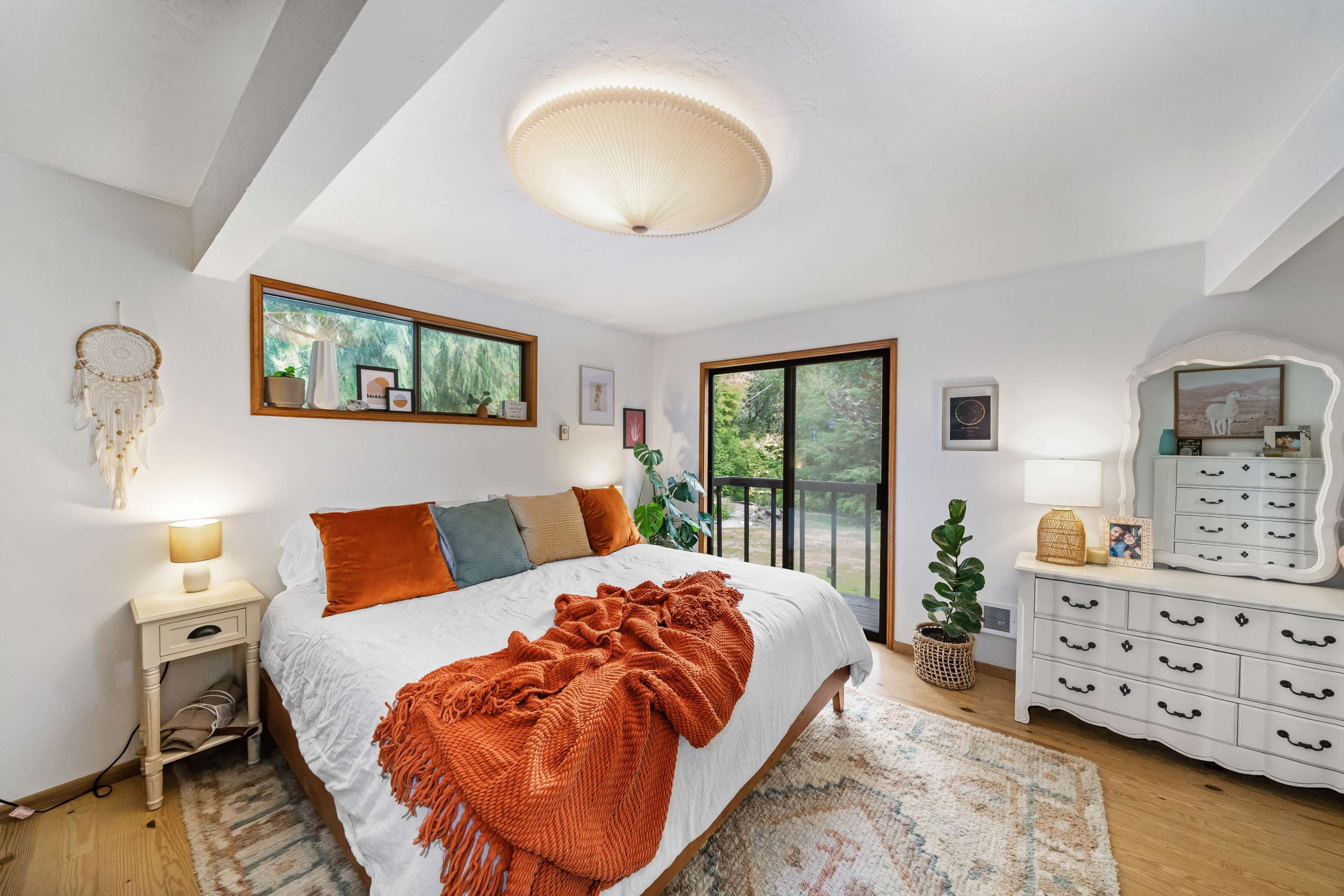 The image shows a cozy bedroom with a large bed, adorned with decorative pillows and a textured blanket, alongside a dresser and large windows overlooking a green exterior.