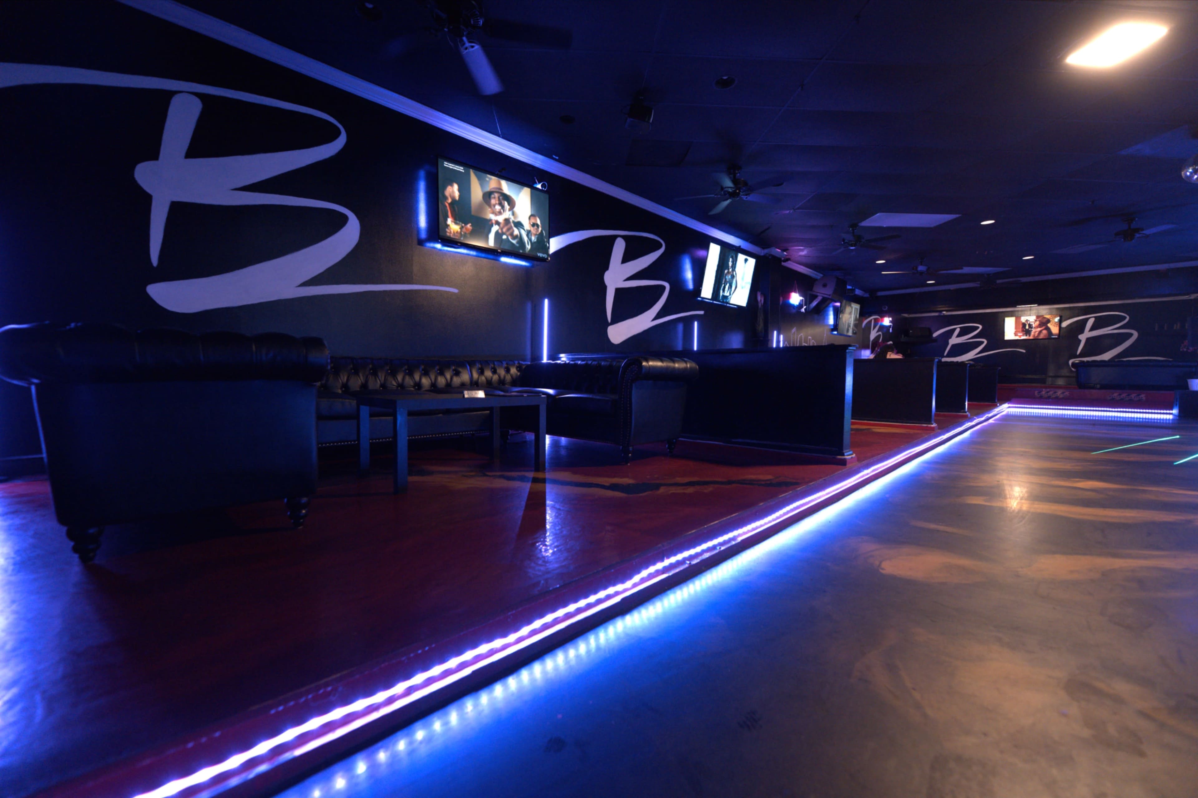 The image shows a dimly lit lounge area with black sofas, illuminated by blue LED lights along the floor and walls, and framed screens displaying images.