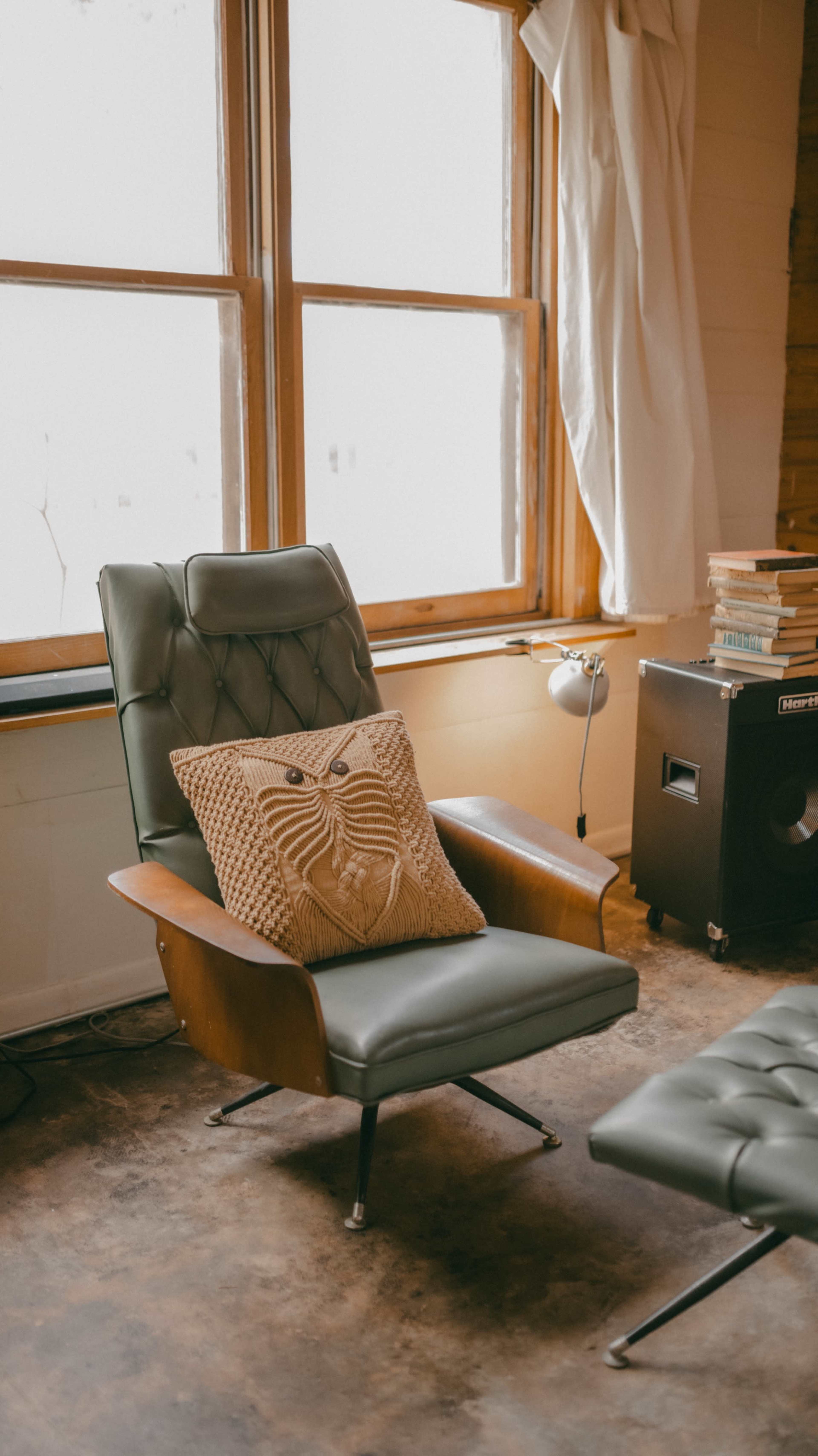 The image shows a modern green armchair with a decorative pillow featuring a face design, positioned near a window in a cozy room with wooden accents.