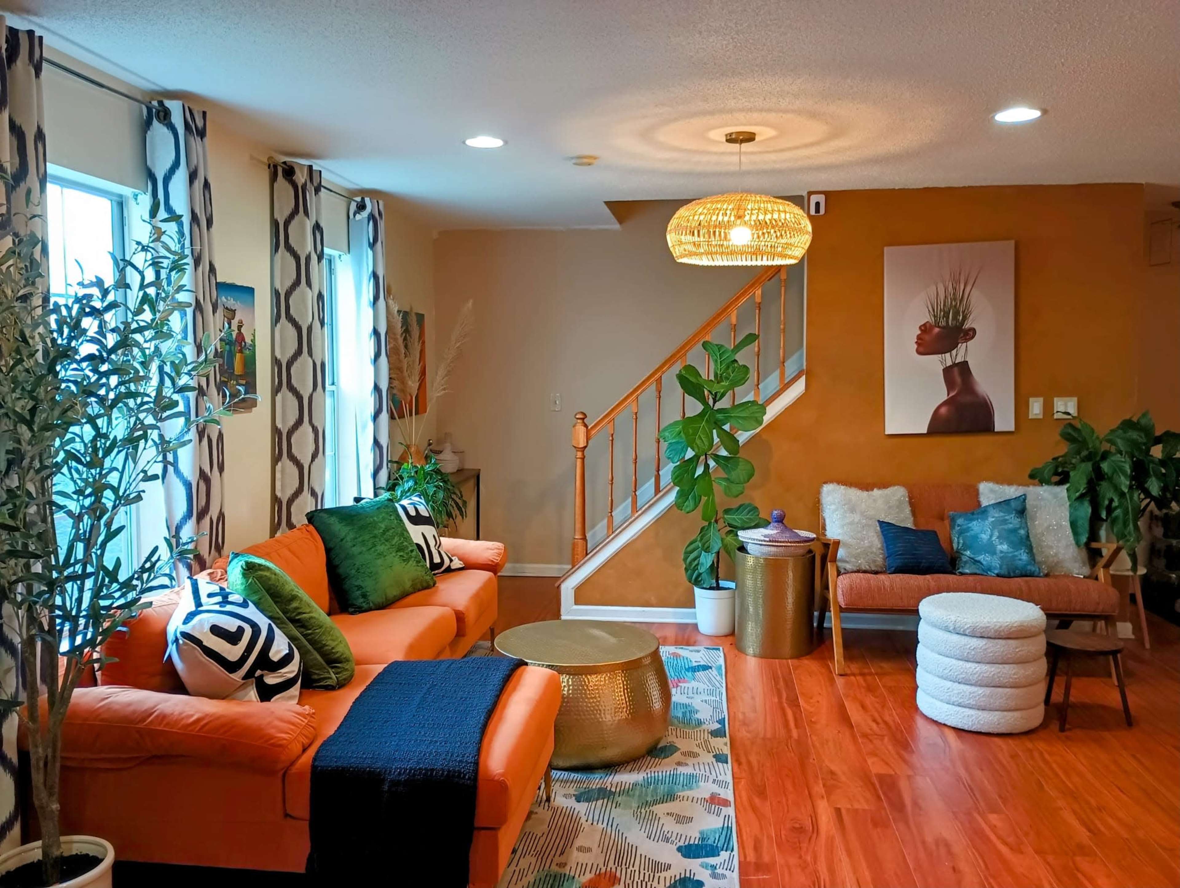 A cozy living room features orange sofas, a round coffee table, and a staircase with a pendant light, surrounded by plants and decorative artwork.
