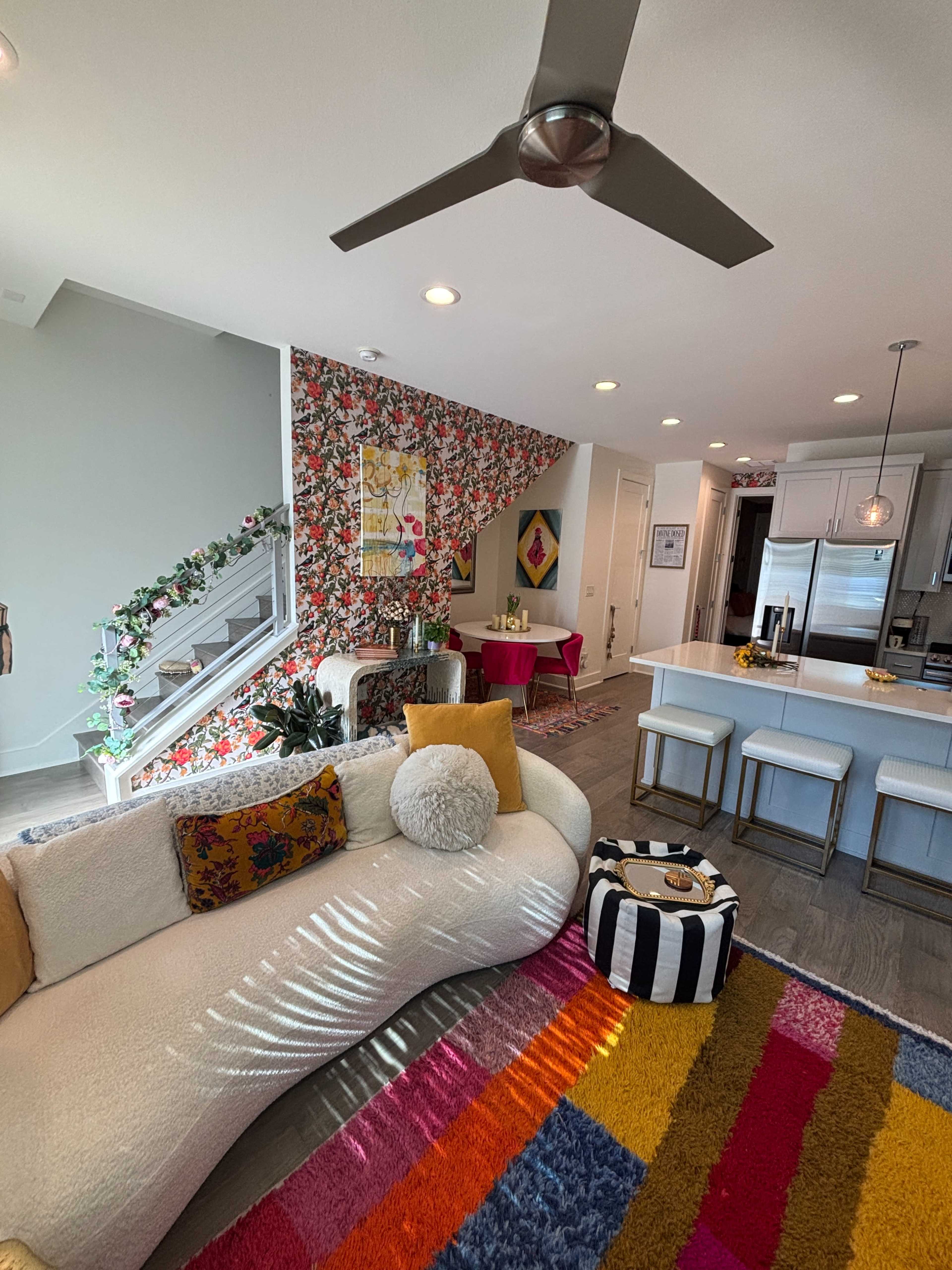 The image features a modern living space with a curved white sofa, a colorful rug, a floral accent wall, and an open kitchen area in the background.
