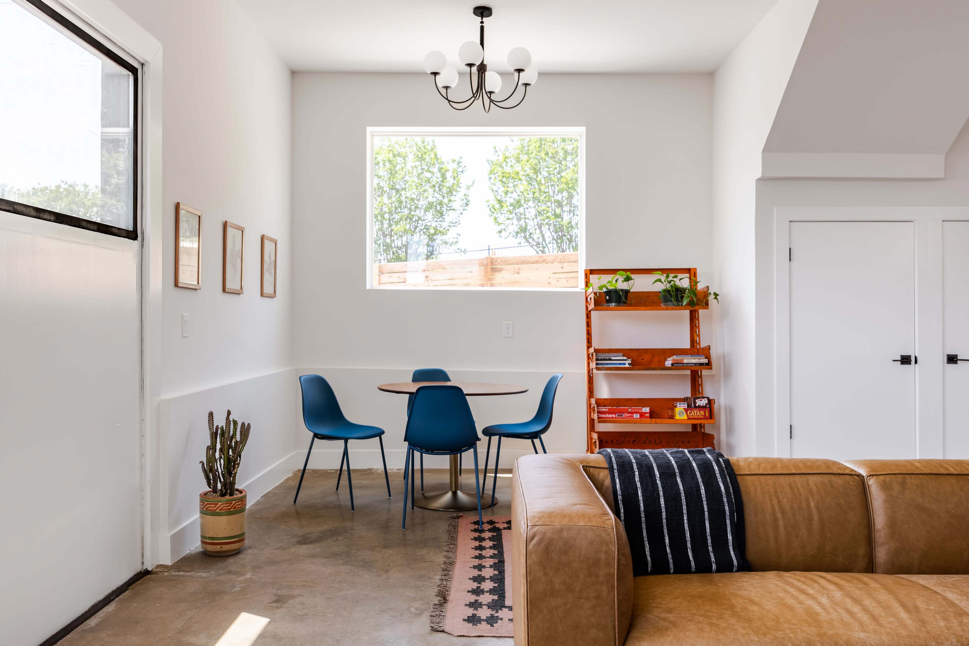 A modern living space featuring a brown leather couch, a round dining table with blue chairs, and a wooden shelving unit with plants and books.