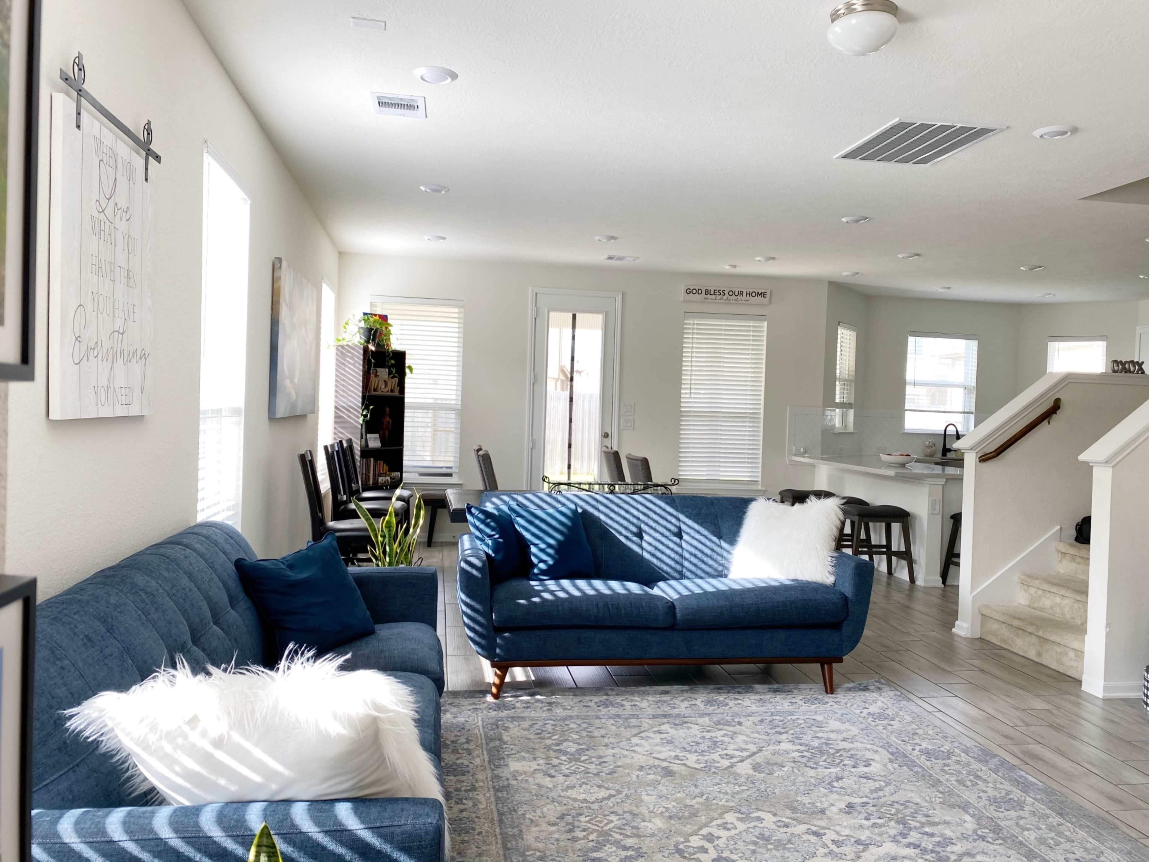 A bright and spacious living room with two blue sofas, a decorative rug, and an open floor plan leading to a dining area and kitchen.