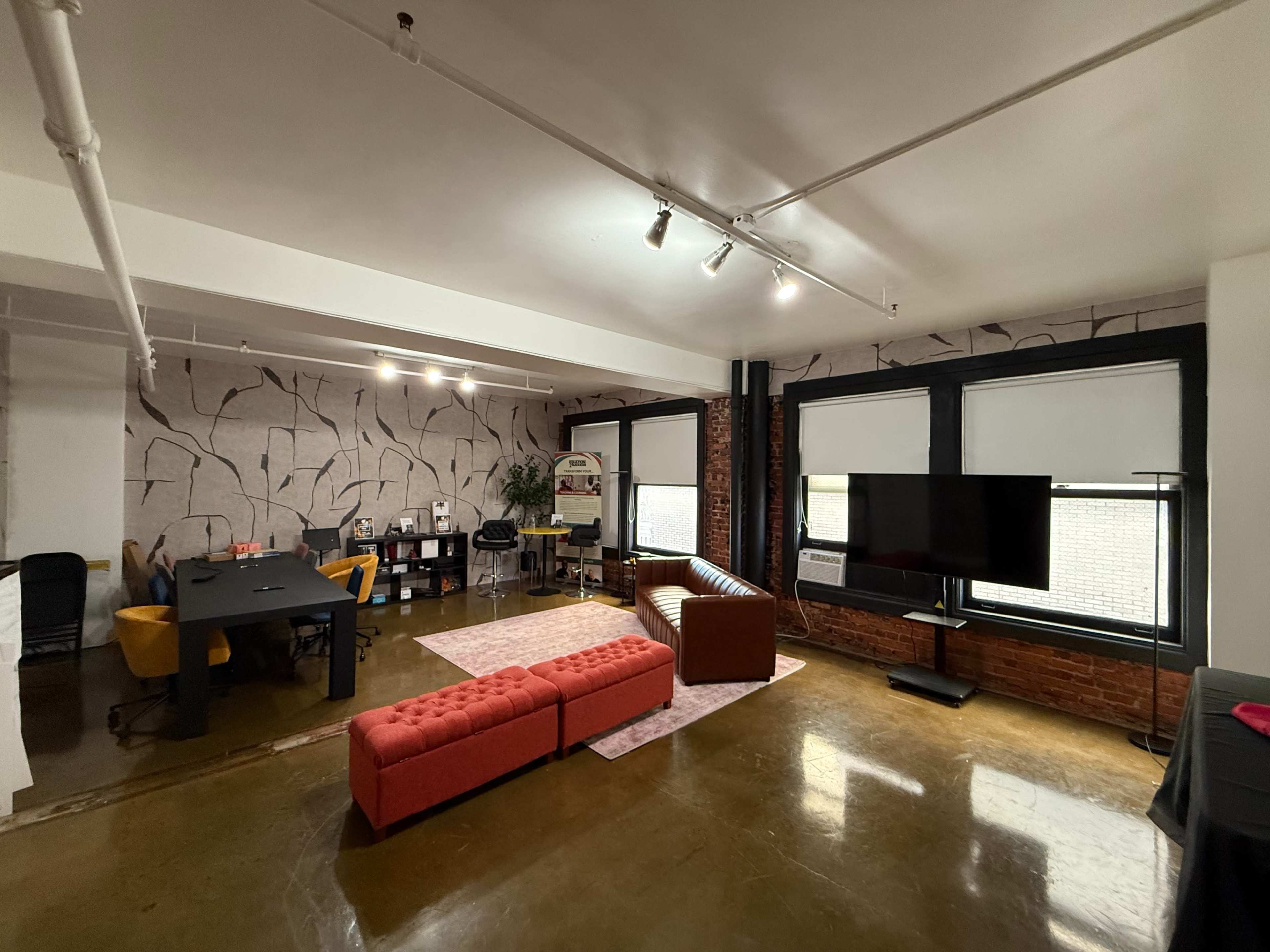 The image shows a modern office space featuring a mix of seating, including a red couch and a brown sofa, a large television, and a workspace with desks and shelves.