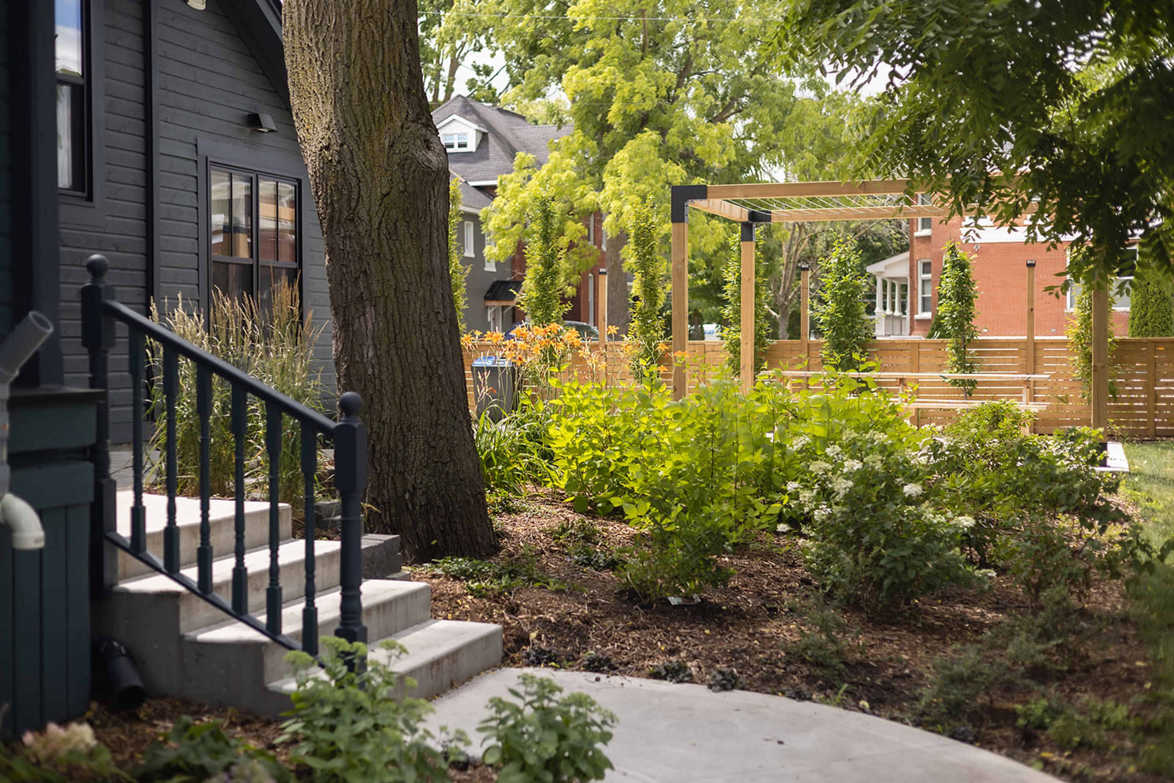 A pathway curves through a landscaped garden featuring greenery and wooden structures beside a dark-colored house.