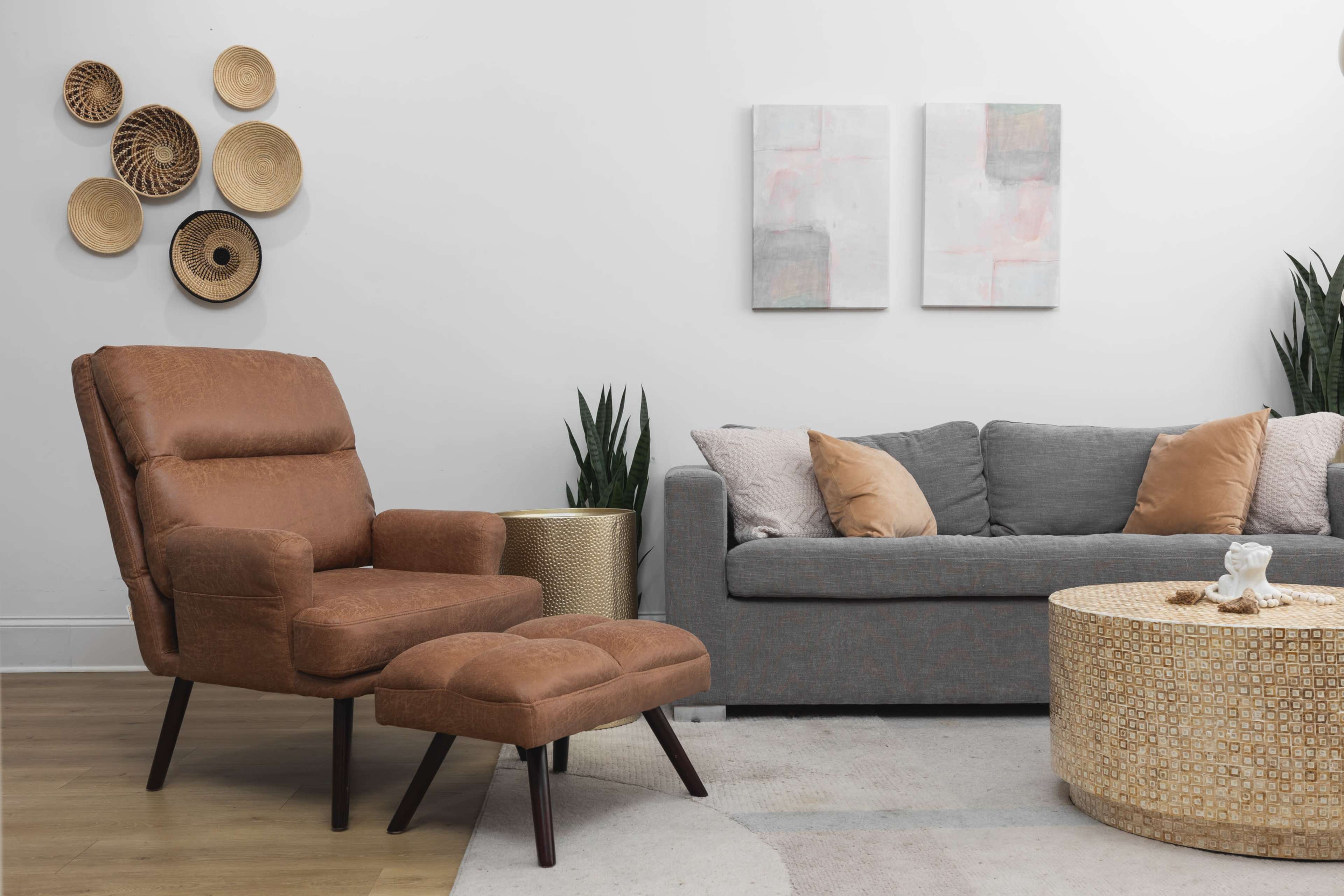 The scene features a cozy living room with a brown upholstered armchair and ottoman, a gray sofa with decorative pillows, a round coffee table, and wall art adorned with woven baskets.