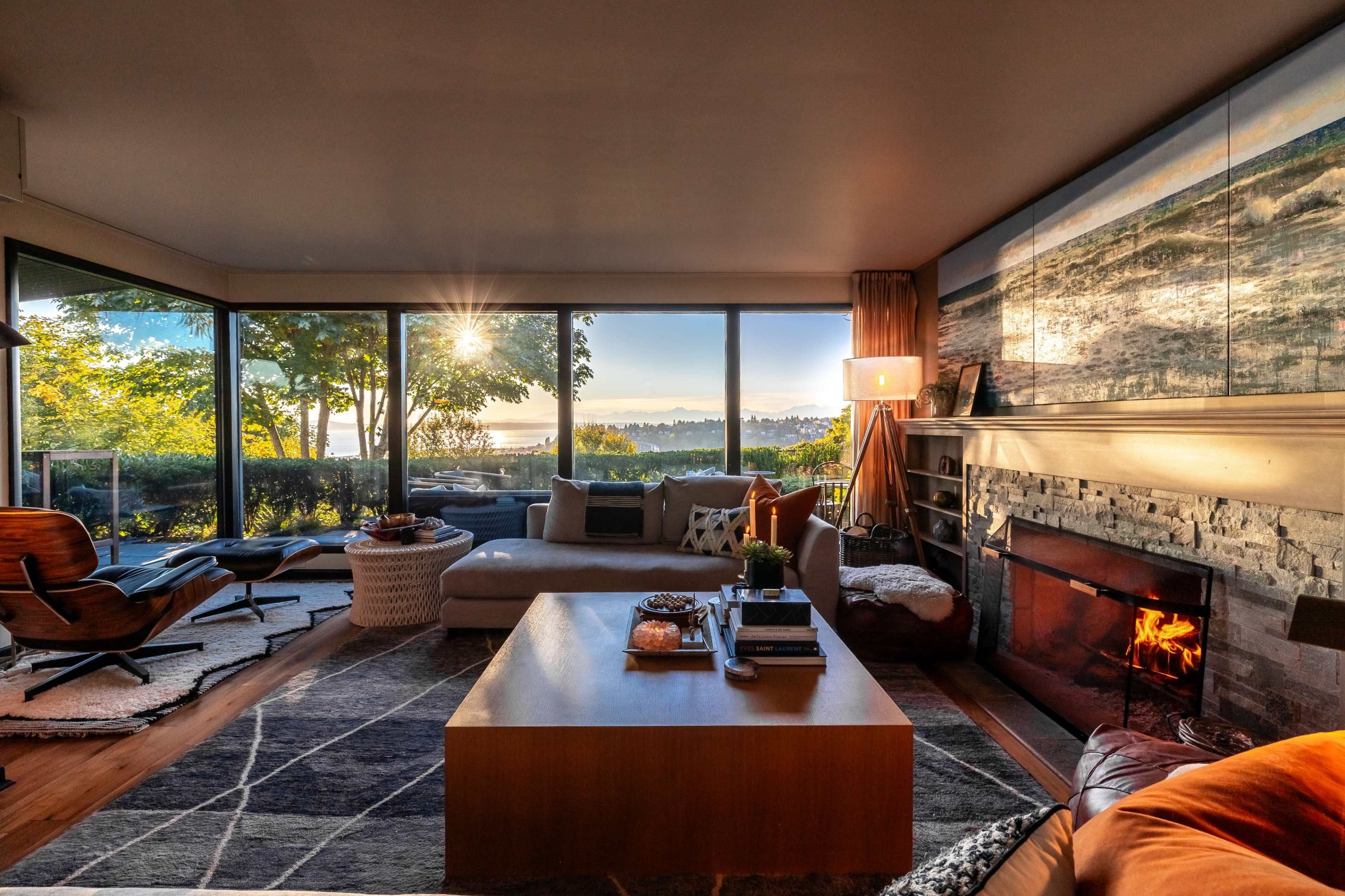 A cozy living room features a fireplace, large windows with a view of trees and a sunset, and modern furniture arranged around a coffee table.