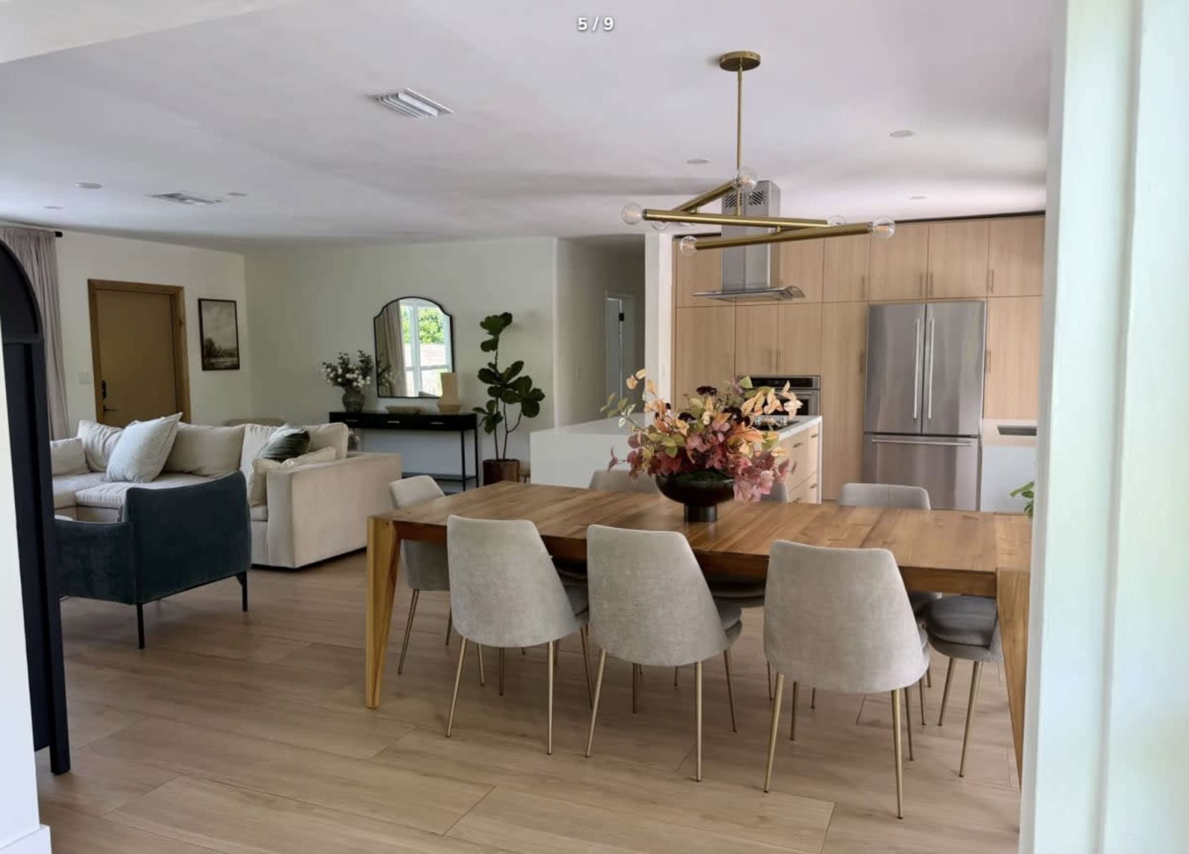The image shows a modern dining area with a wooden table surrounded by beige chairs, adjacent to a kitchen with wooden cabinetry and a living area featuring a light-colored sofa.