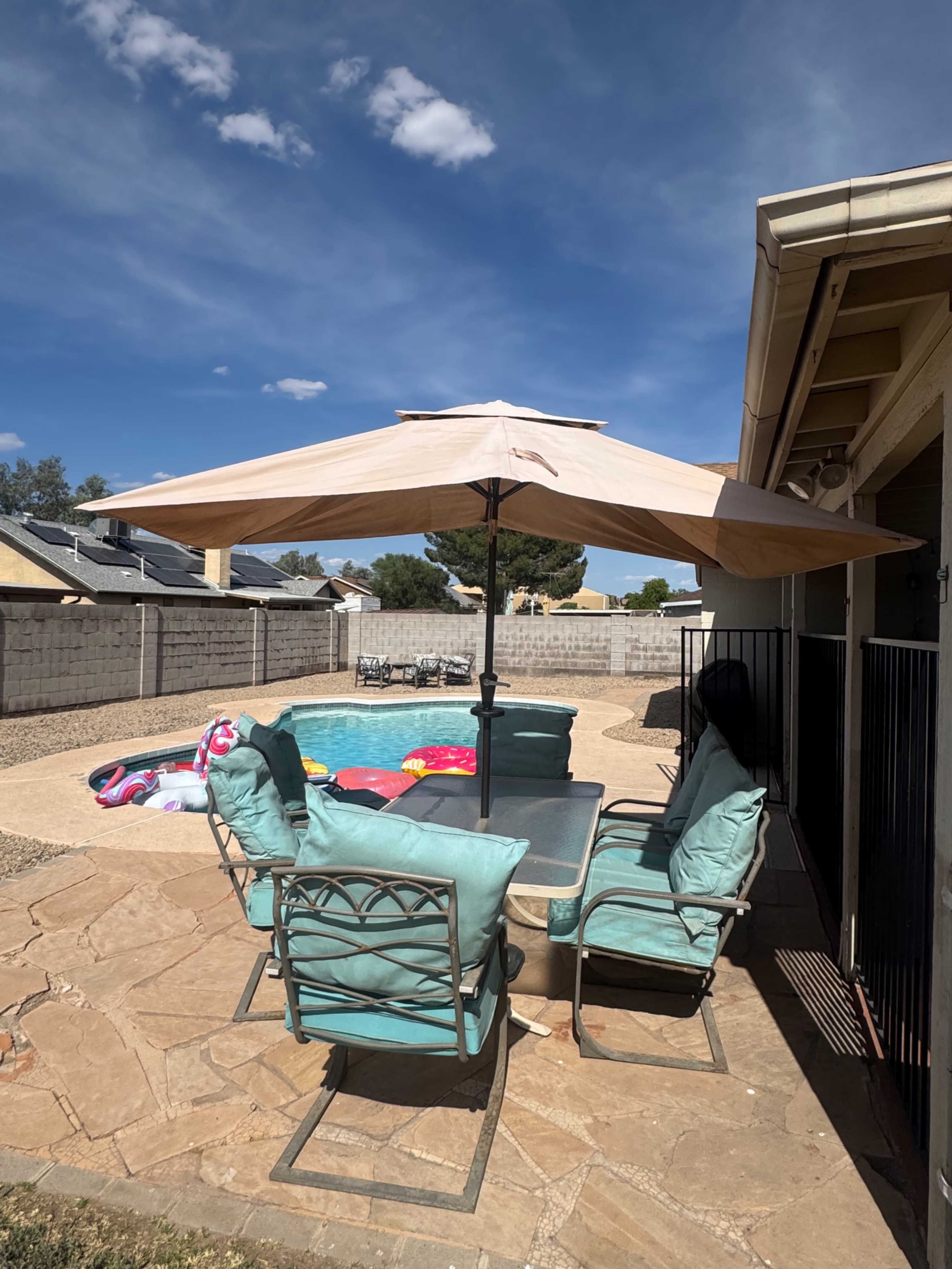 A patio with a table and chairs is shaded by a large umbrella, overlooking a swimming pool surrounded by a stone pathway and a wall in the background.