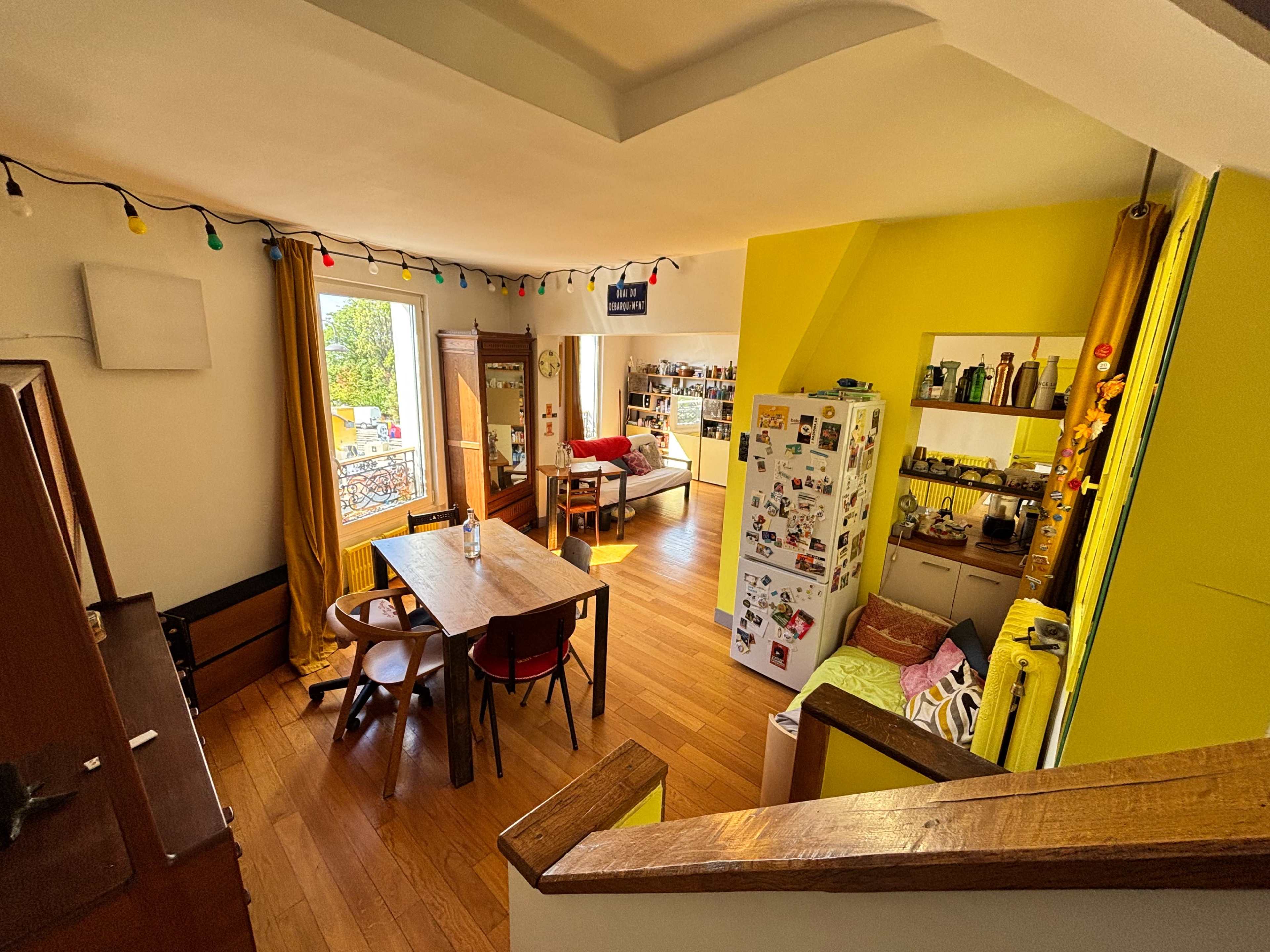 The image shows a bright, open kitchen and living area with wooden flooring, yellow walls, and a dining table surrounded by chairs, along with decorative string lights.