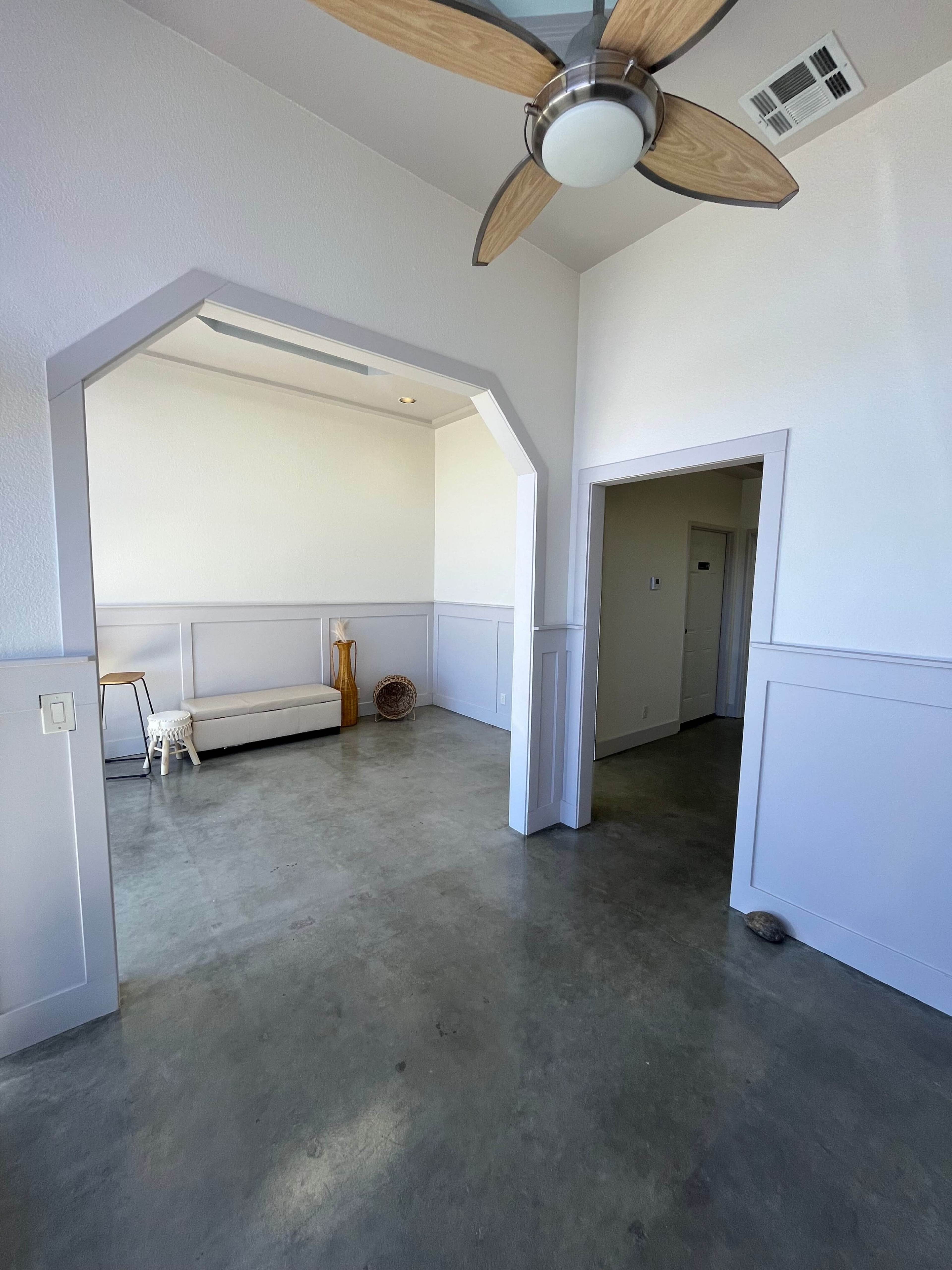 The image shows a light-filled room with a concrete floor, featuring an arched entryway, a seating bench, decorative items, and a door leading to another area.