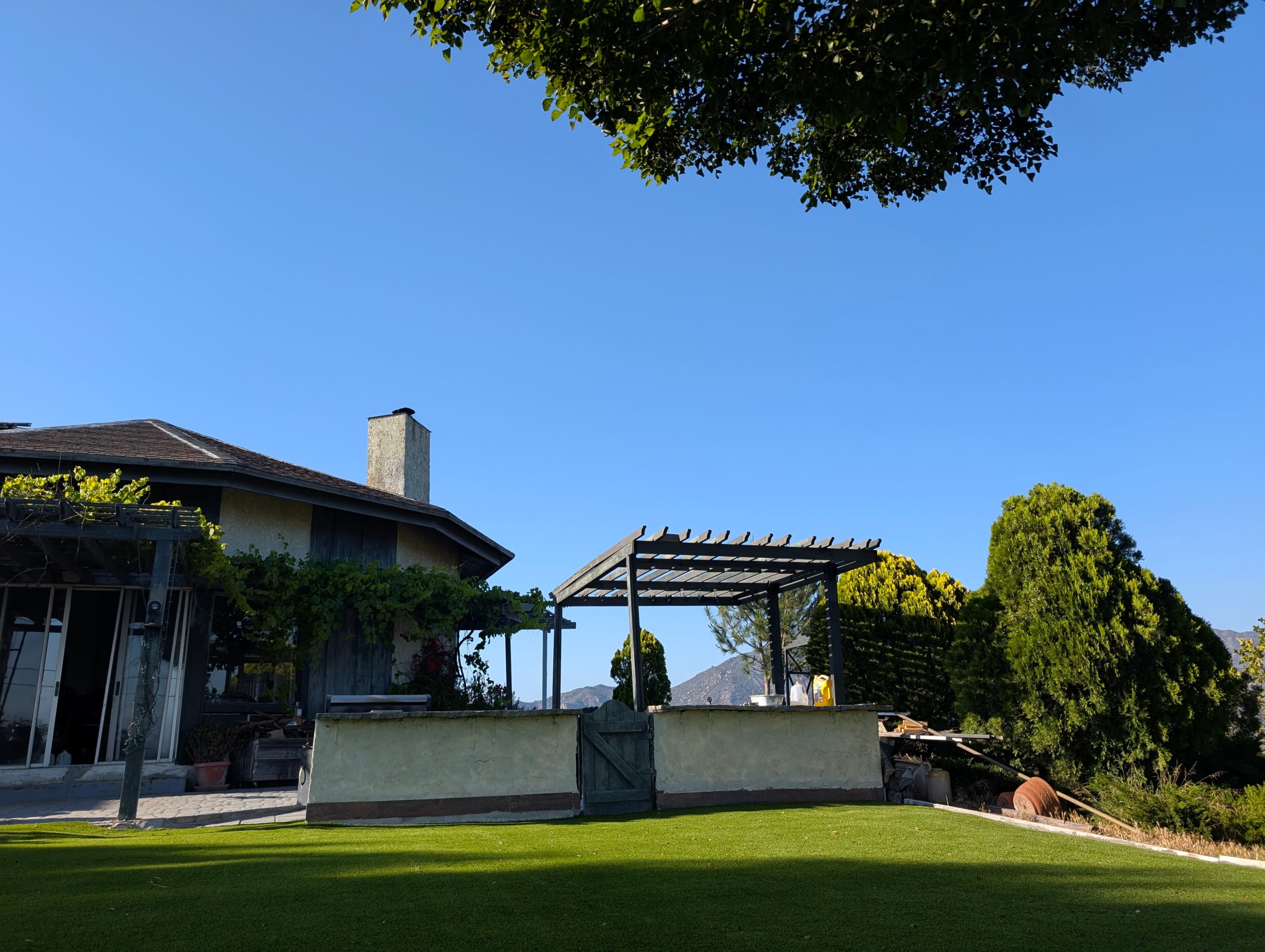 A house with a wooden pergola and a lush green lawn is set against a clear blue sky and distant mountains.