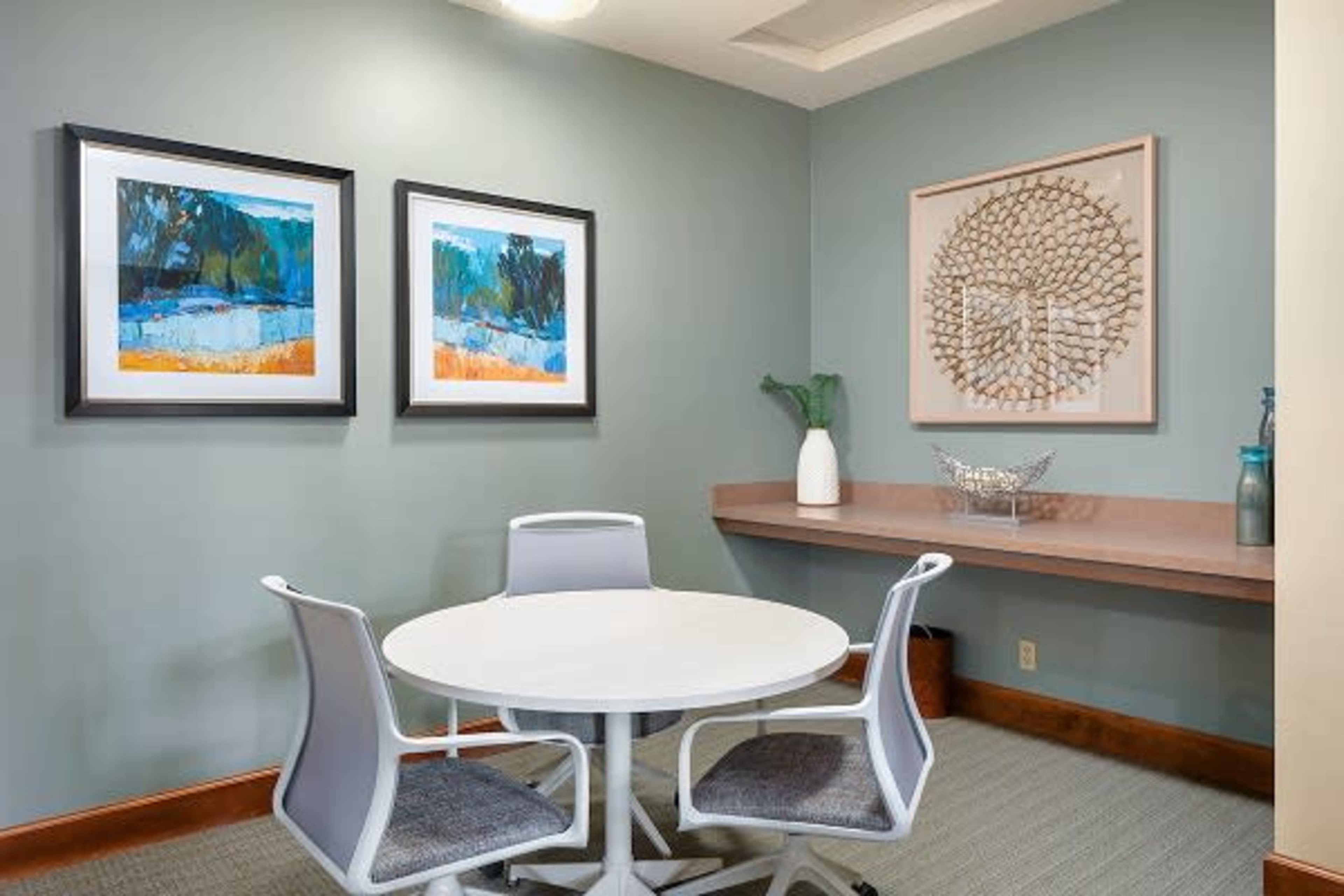 A small meeting room featuring a round white table surrounded by three modern chairs and decorated with framed artworks on the walls.