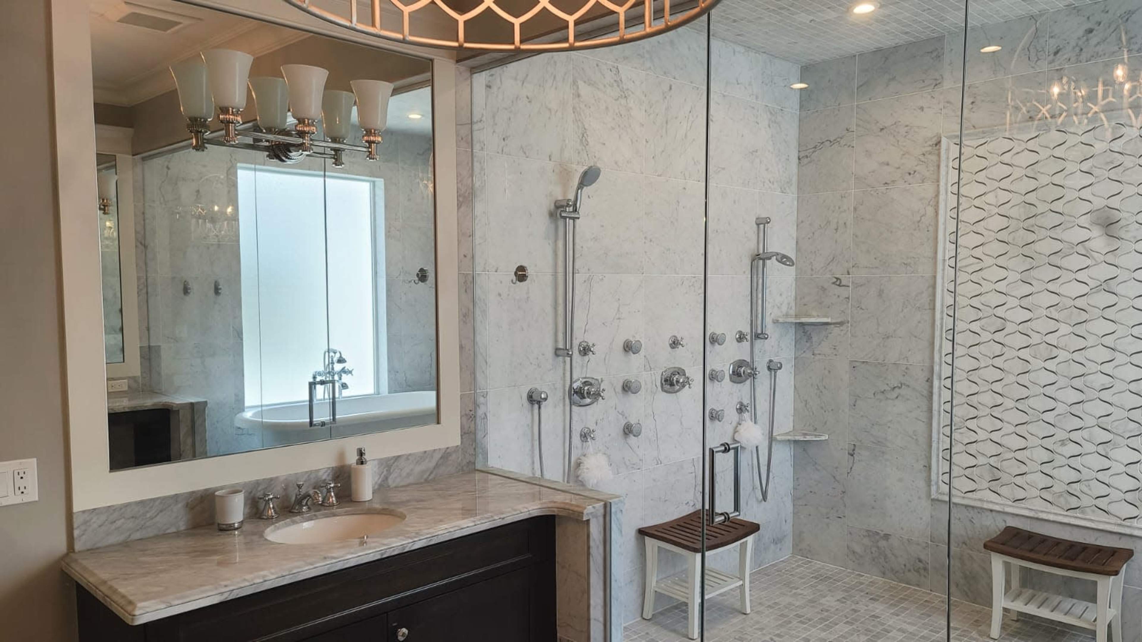 A modern bathroom featuring a marble shower with multiple showerheads, a freestanding bathtub, and a mirrored vanity with four light fixtures.