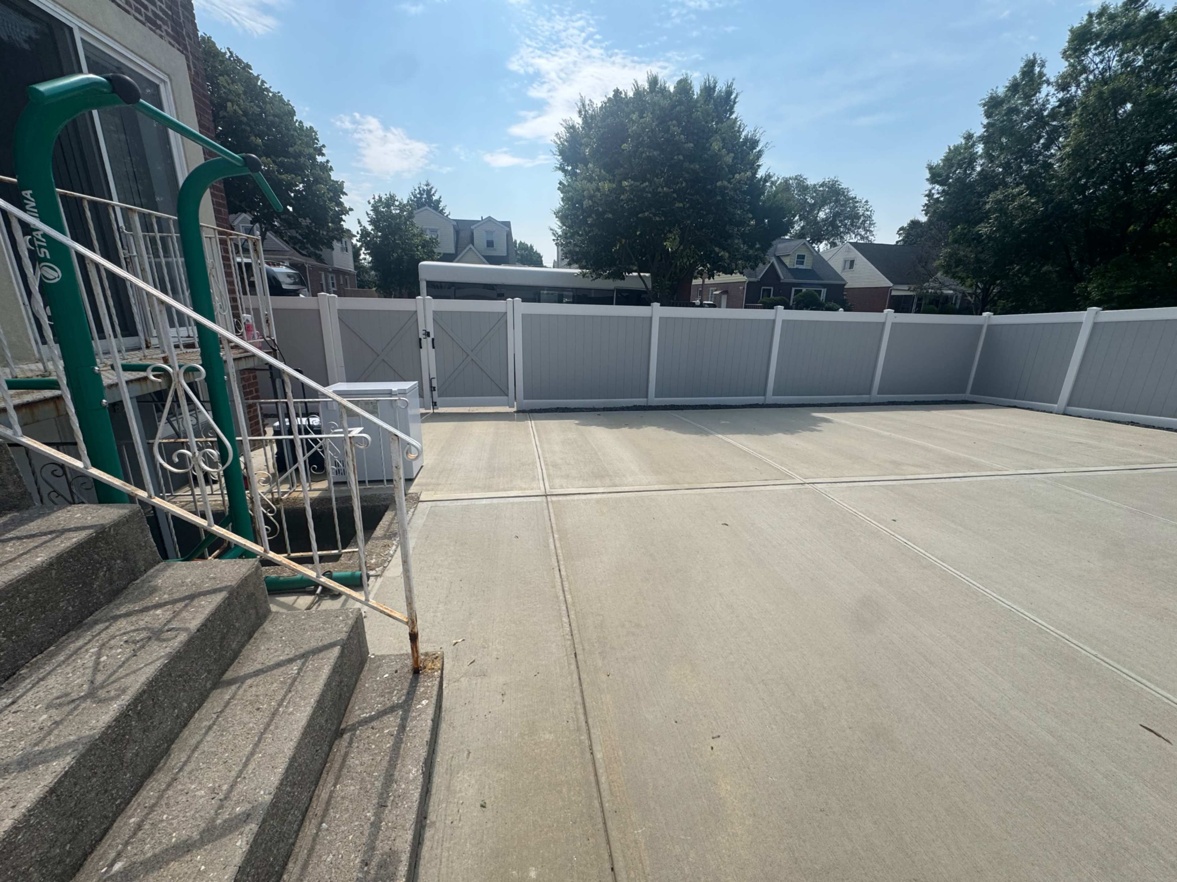 The image shows a concrete area next to a set of stairs with a green railing, enclosed by a white fence and surrounded by trees and houses in the background.