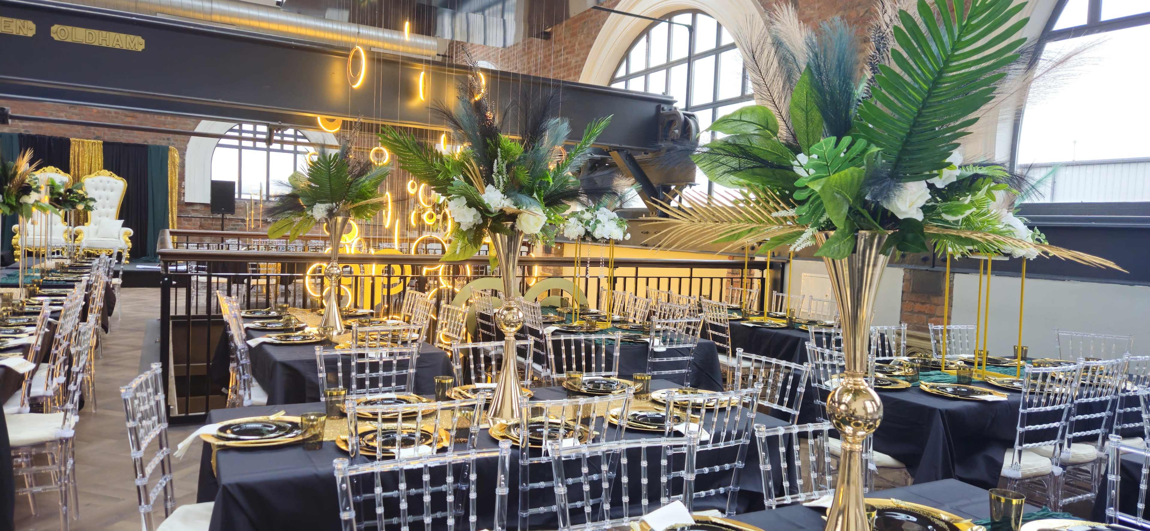 A large dining area is arranged with elegant tables set with black tablecloths, gold accents, and tall centerpieces featuring greenery and white flowers.