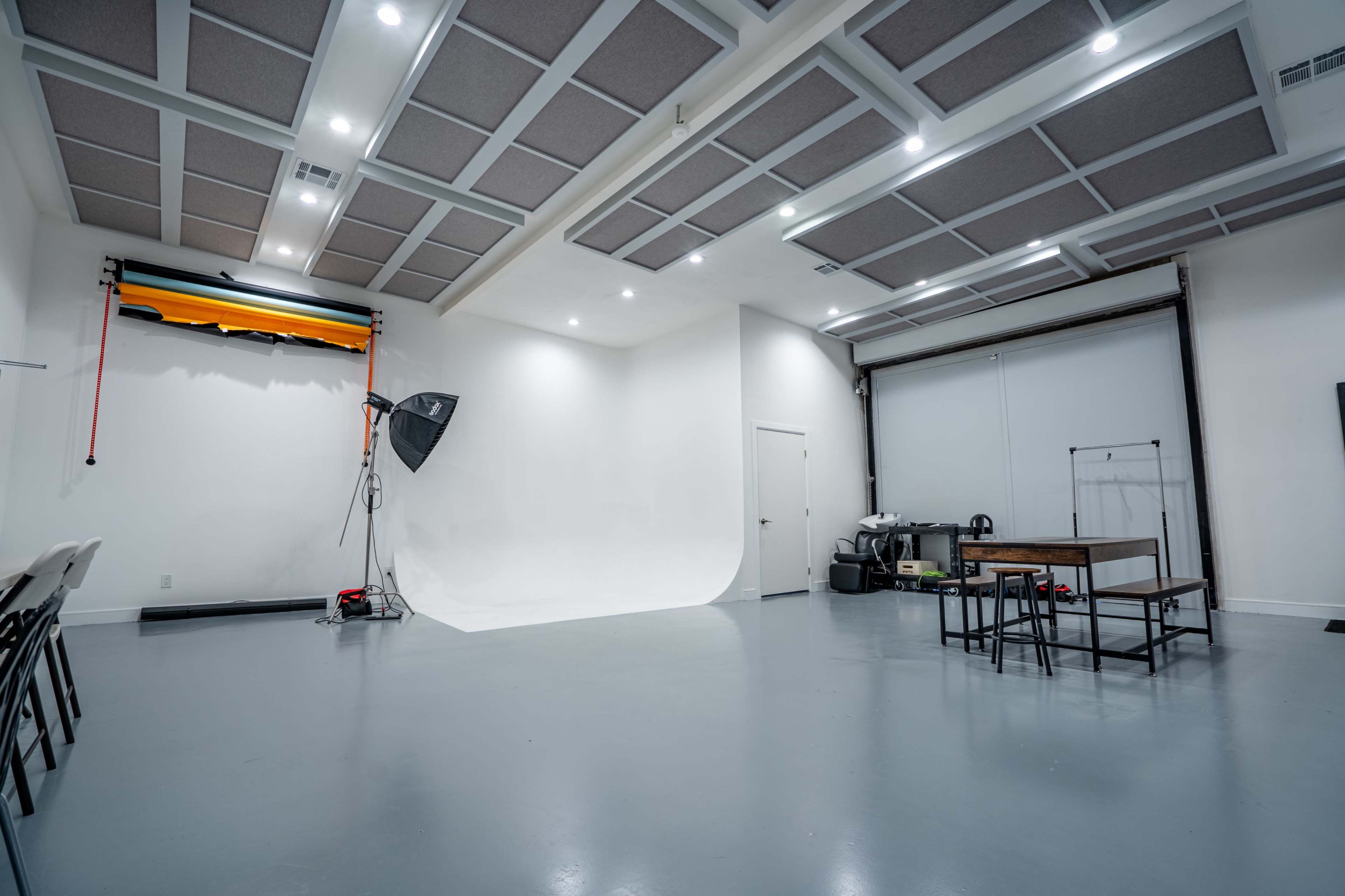 The image shows a spacious photo studio with a white backdrop, soft lighting, and a few pieces of equipment, including a backdrop holder and a table.