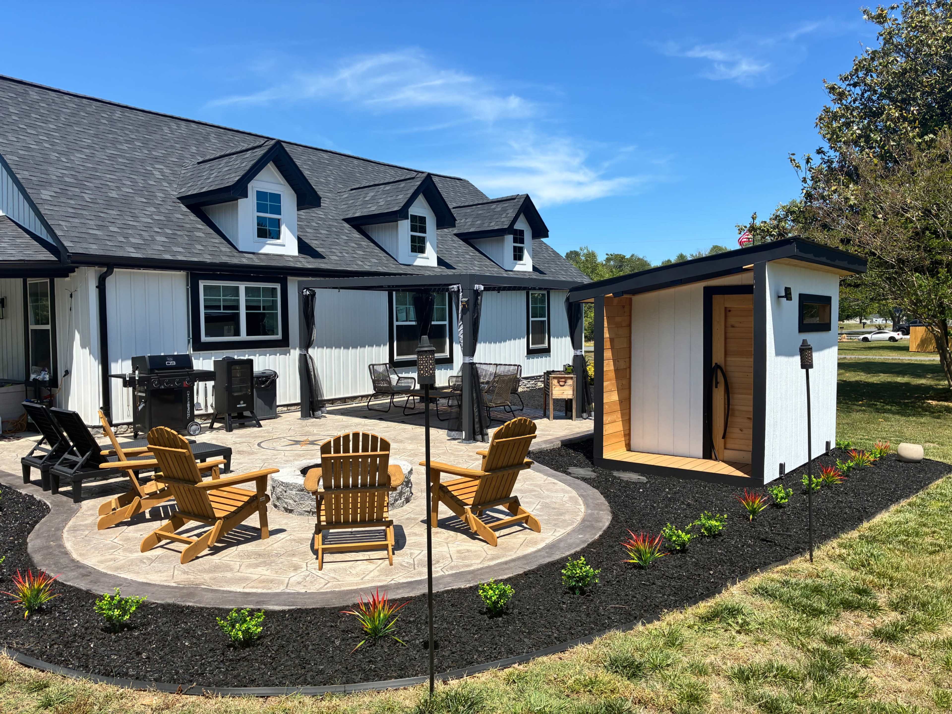 A circular stone patio with wooden Adirondack chairs is surrounded by landscaping and a small structure in a backyard next to a house.