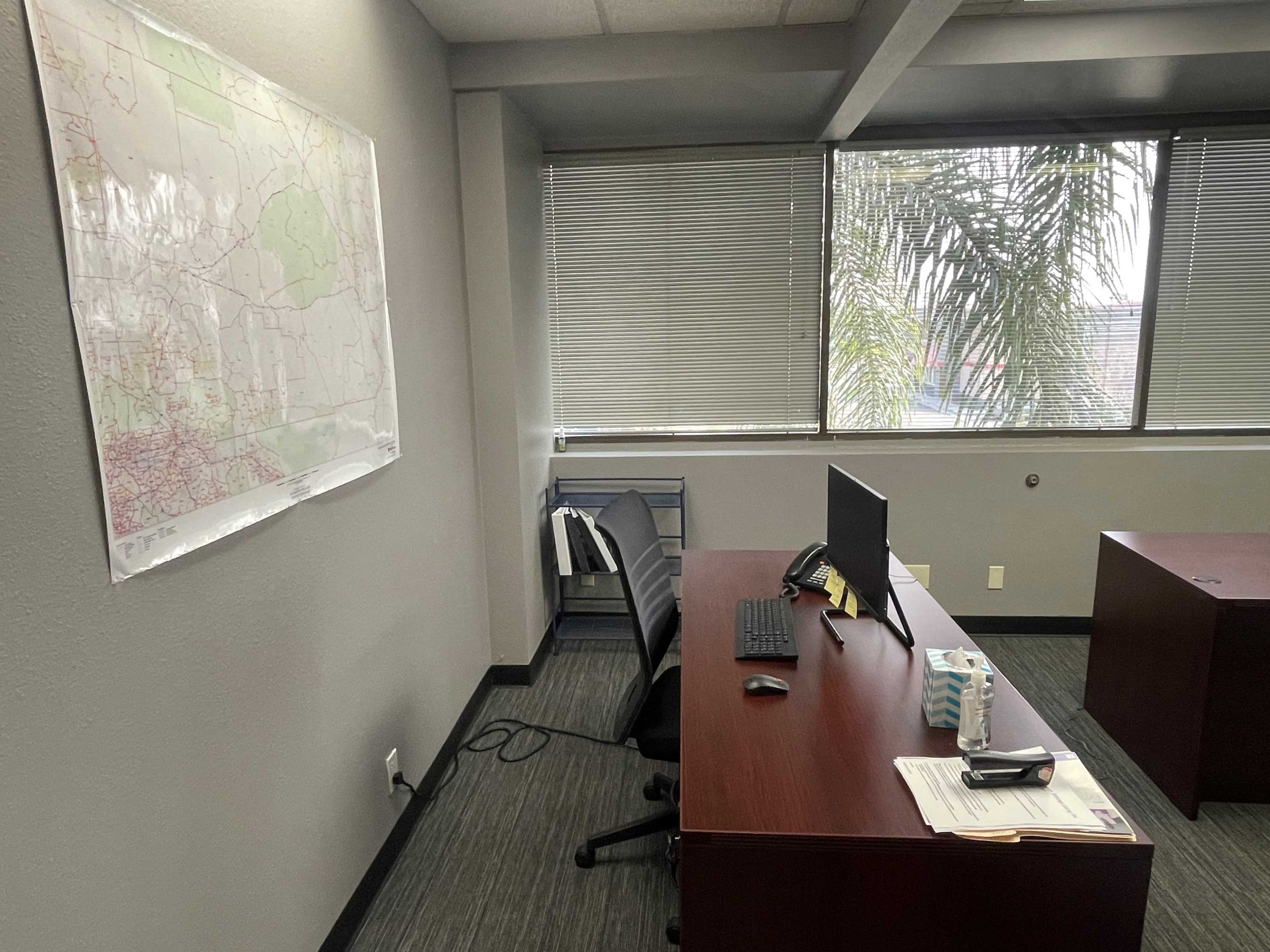 An office space featuring two wooden desks, a computer on one desk, a map on the wall, and a window with blinds allowing some natural light.