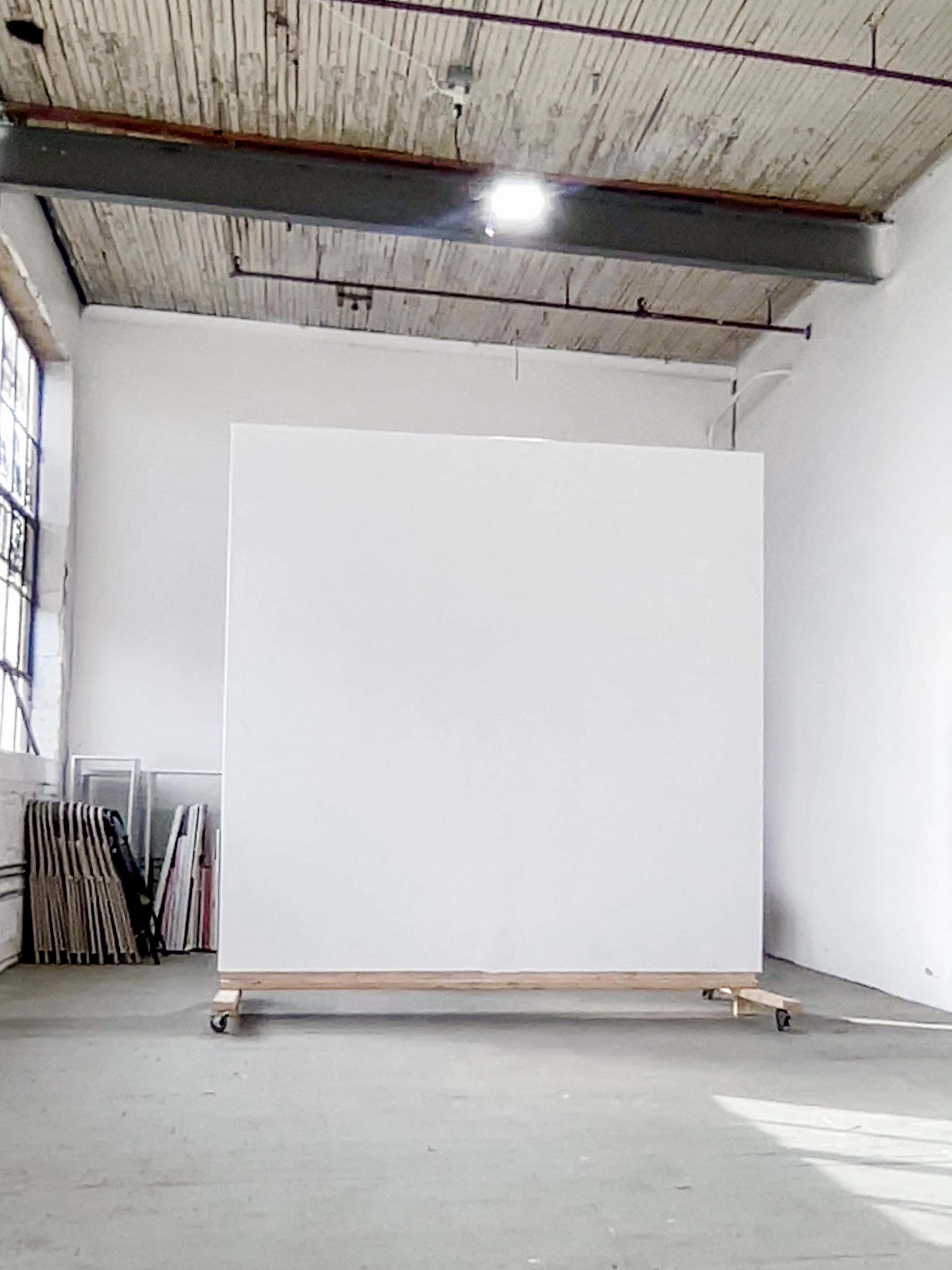 The image shows a large, empty white canvas on wheels in an industrial-style art studio.