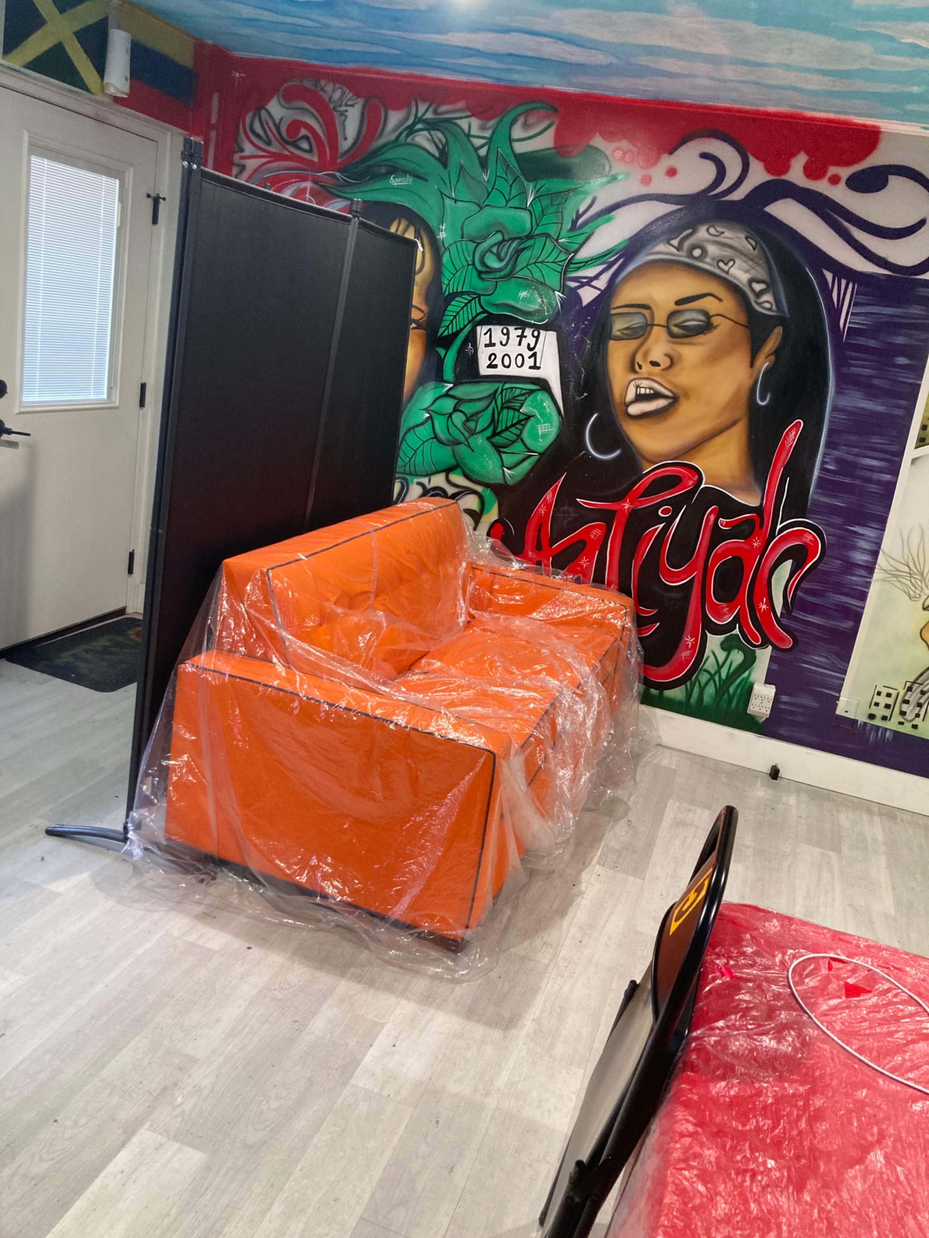 A bright orange sofa wrapped in plastic is positioned next to a partition in a room featuring a colorful mural on the wall.