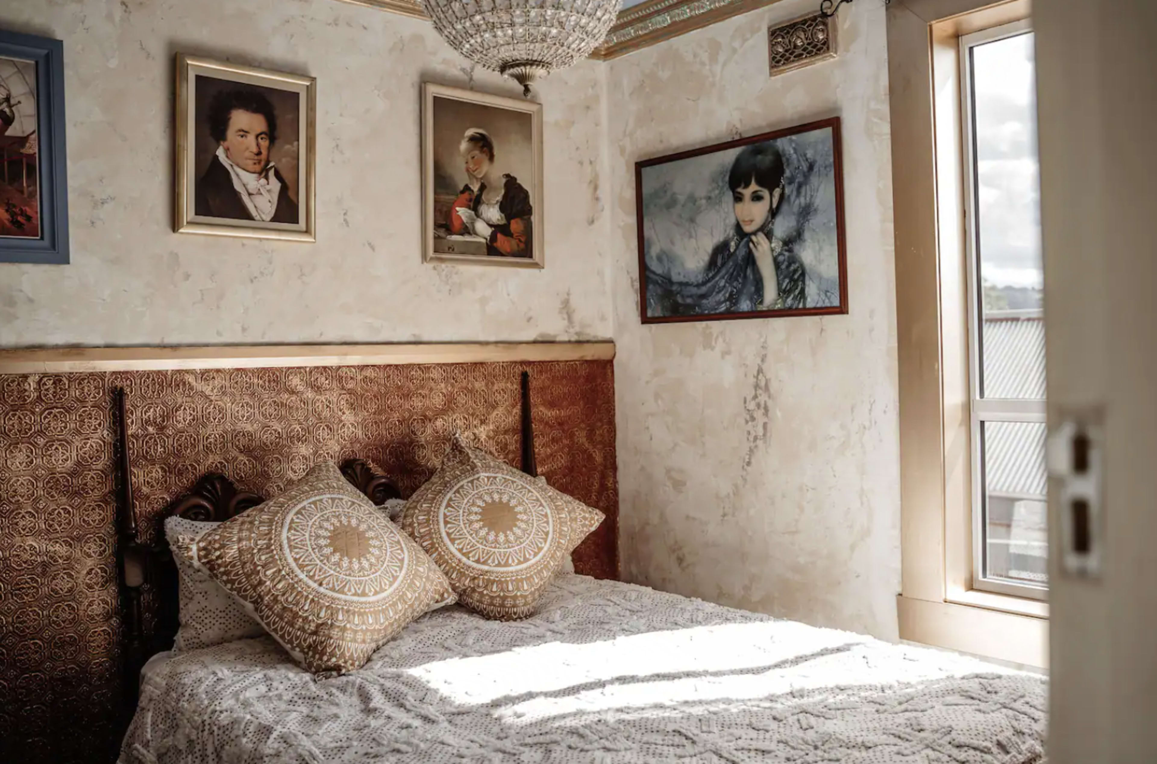 The image shows a bedroom with a bed adorned with decorative cushions, framed portraits on the walls, and a chandelier hanging from the ceiling.