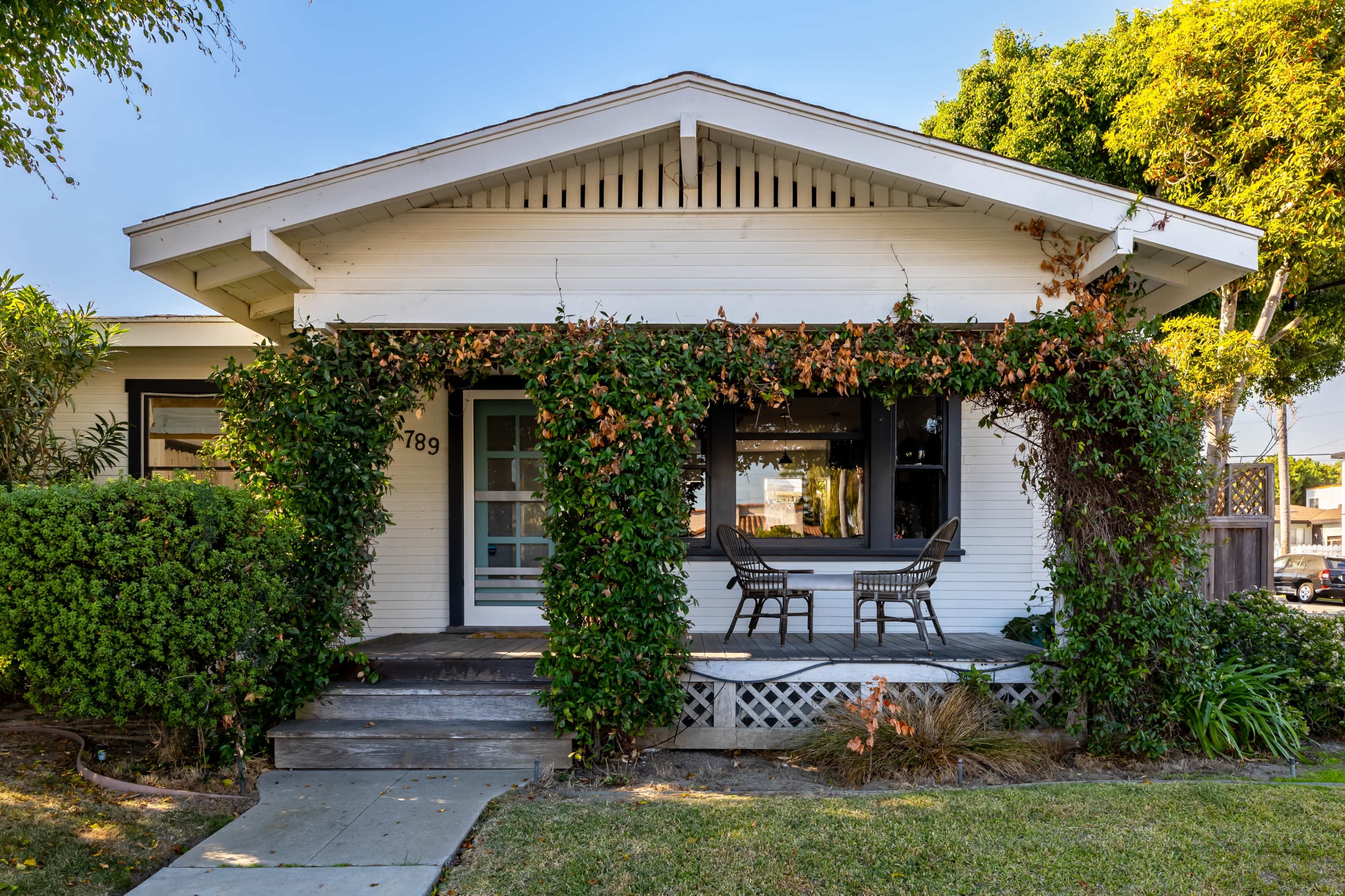 A white house with a front porch is surrounded by greenery and features two chairs on the porch.