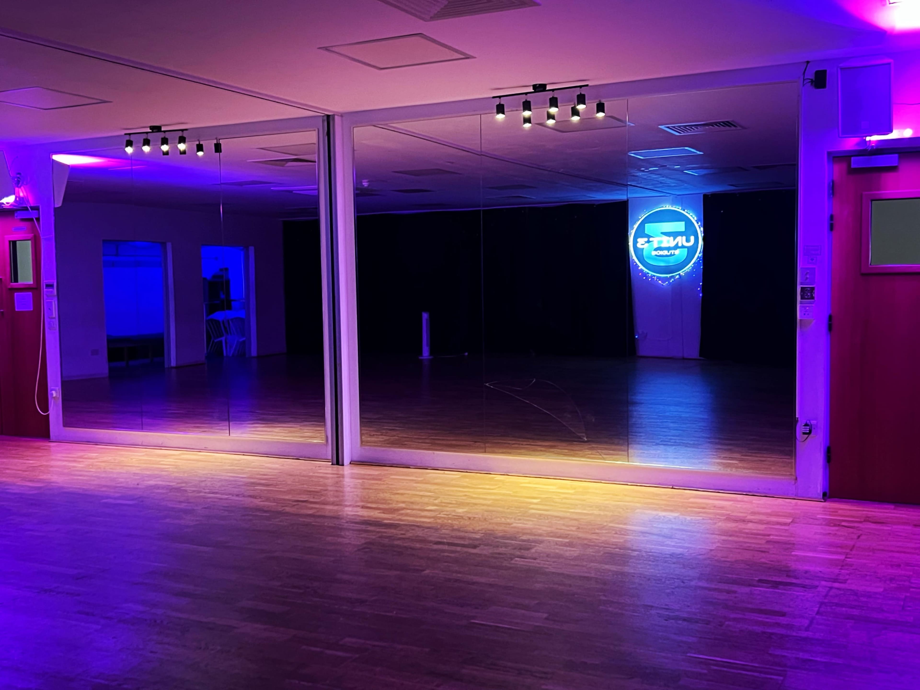 The image shows a spacious dance studio with mirrored walls and colorful lighting, reflecting a neon sign in the background.