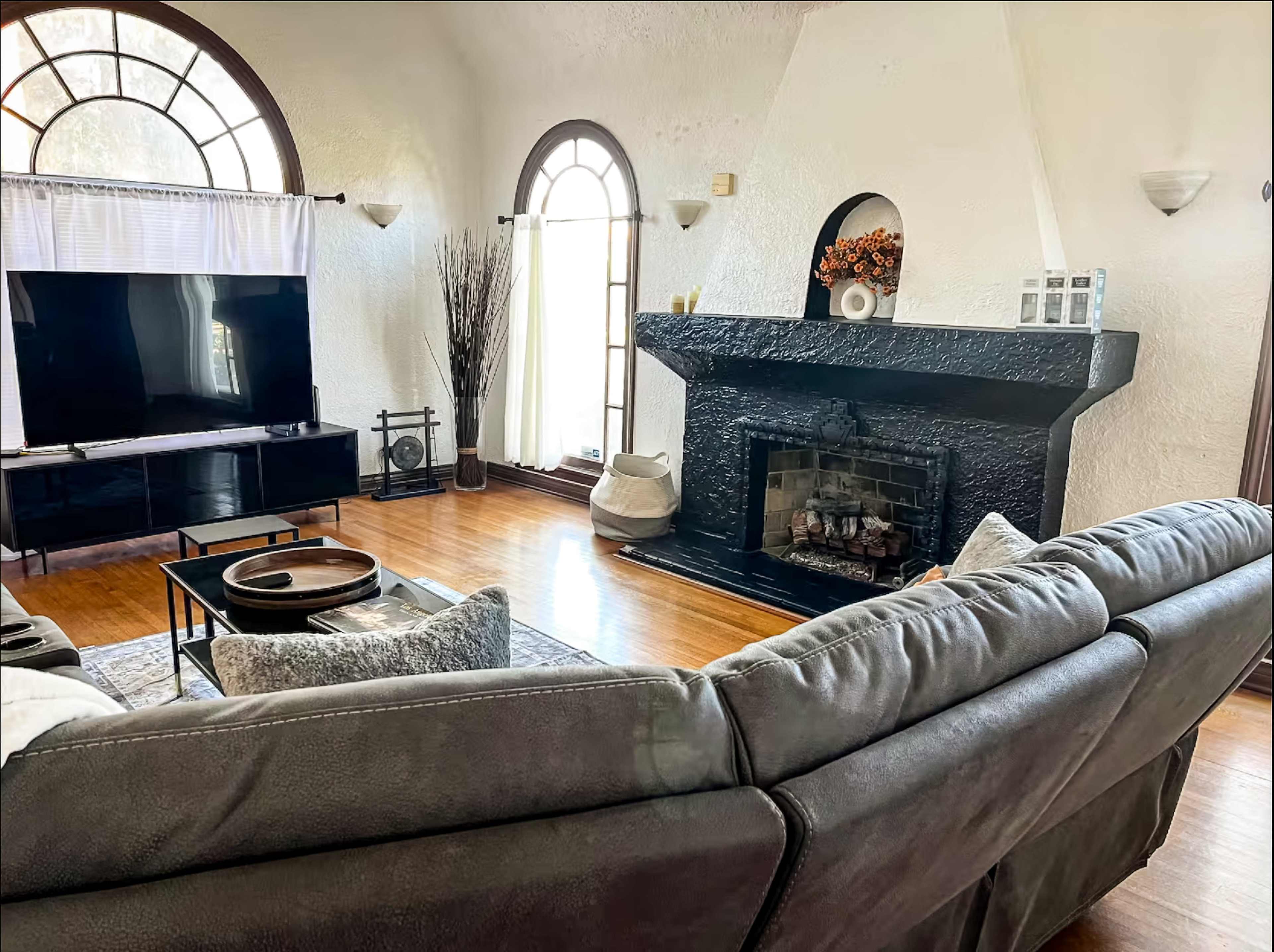 The image shows a living room with a dark stone fireplace, large windows, a television unit, and a comfortable sectional sofa facing the fireplace.