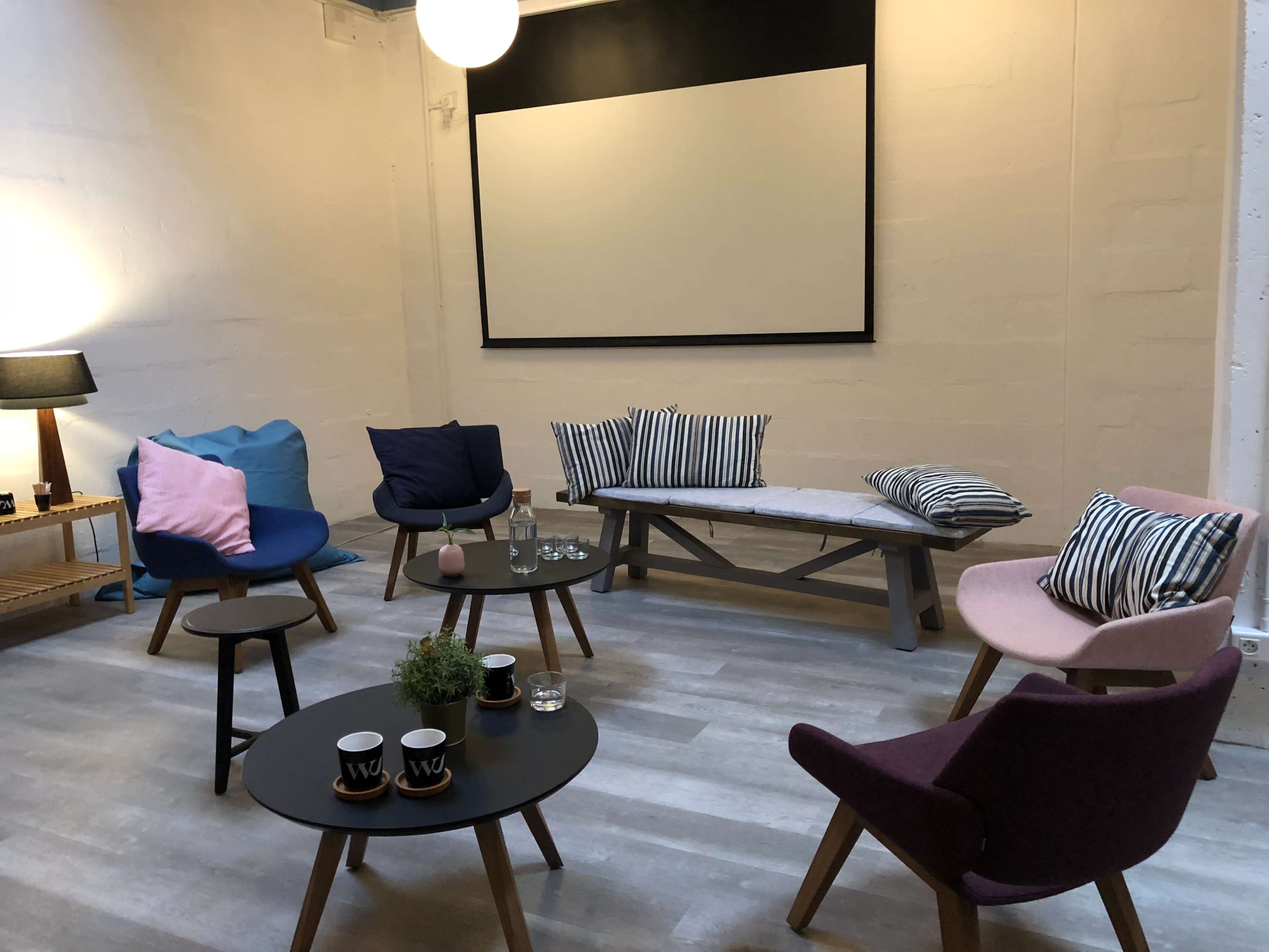 The scene shows a cozy lounge area with various chairs, a bench, tables, and a projector screen at the front.