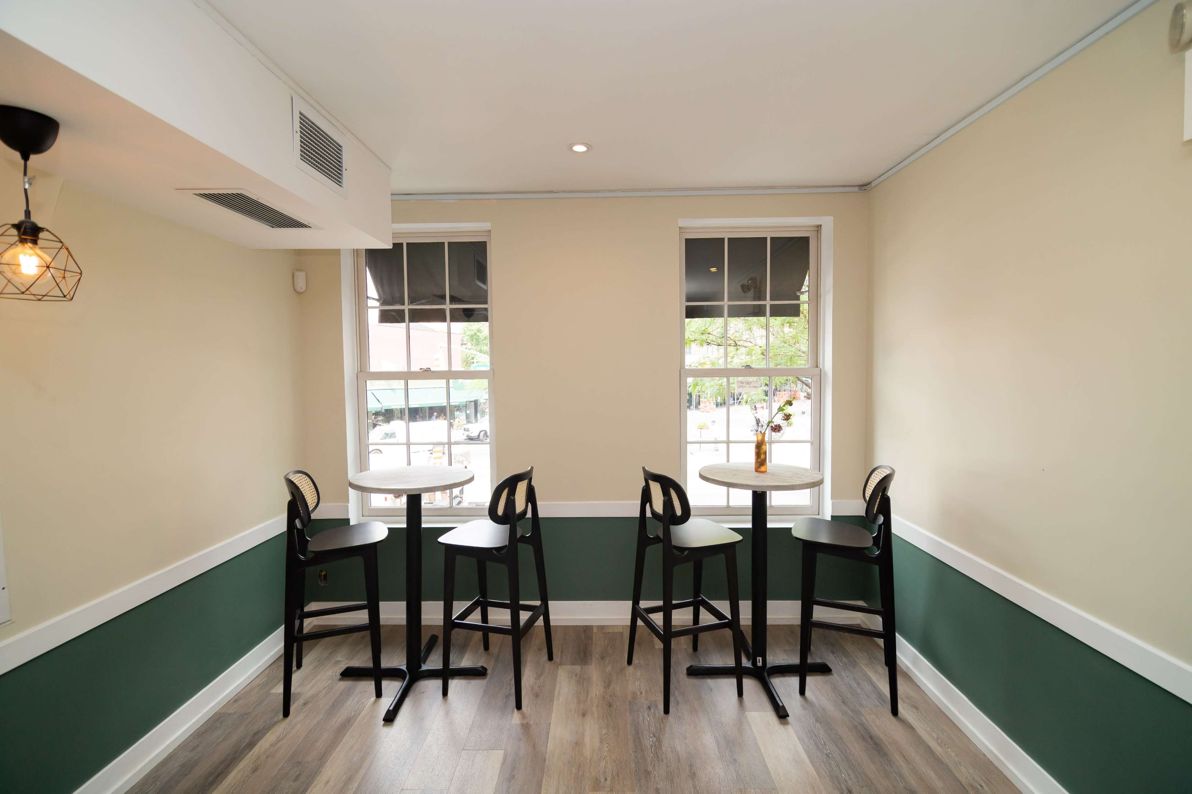 The image shows a small, minimally decorated café space with four black high chairs and tables near large windows.
