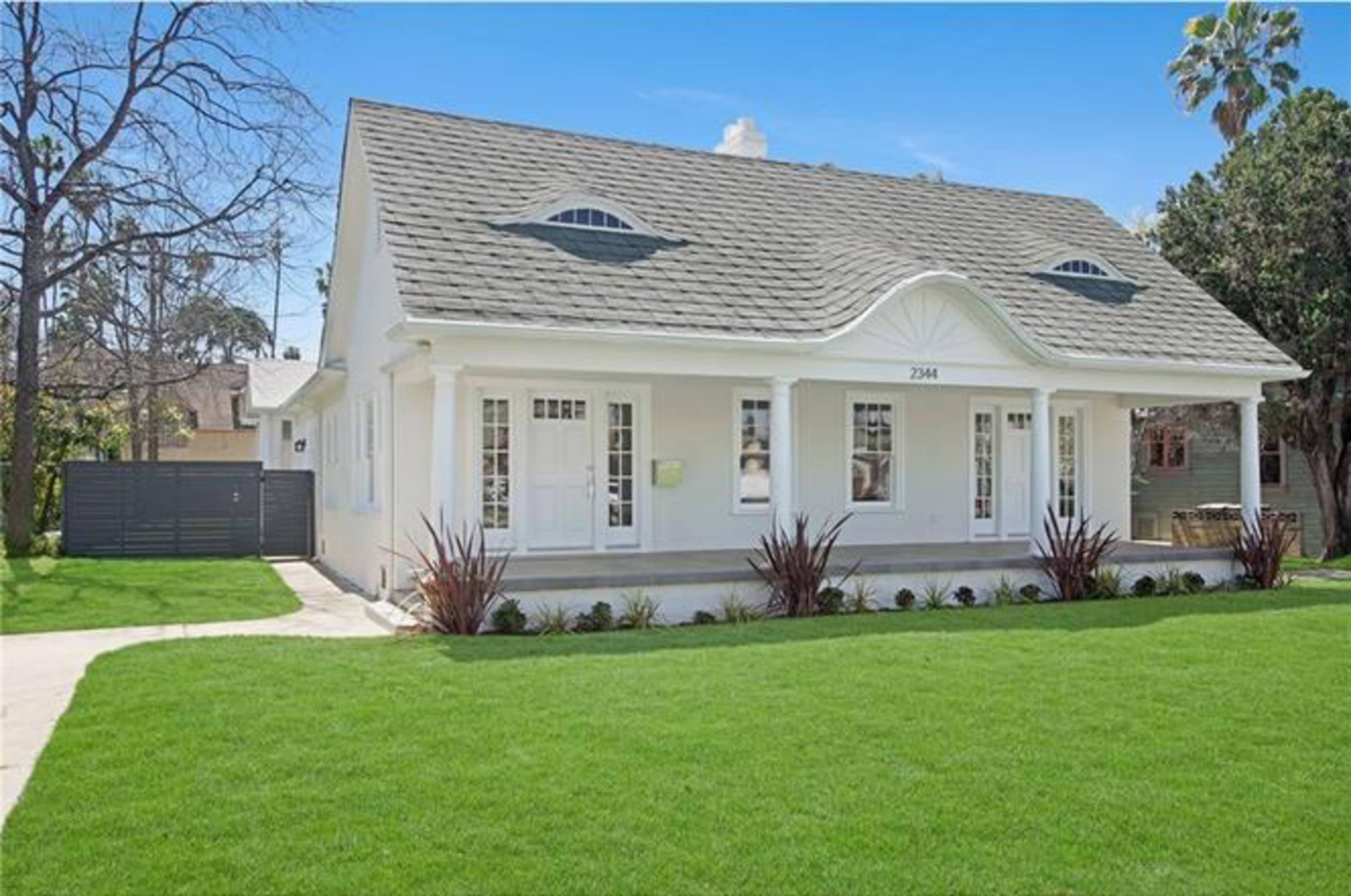 A white, single-story house with a triangular roof and decorative front entry is surrounded by well-maintained grass and landscaping.