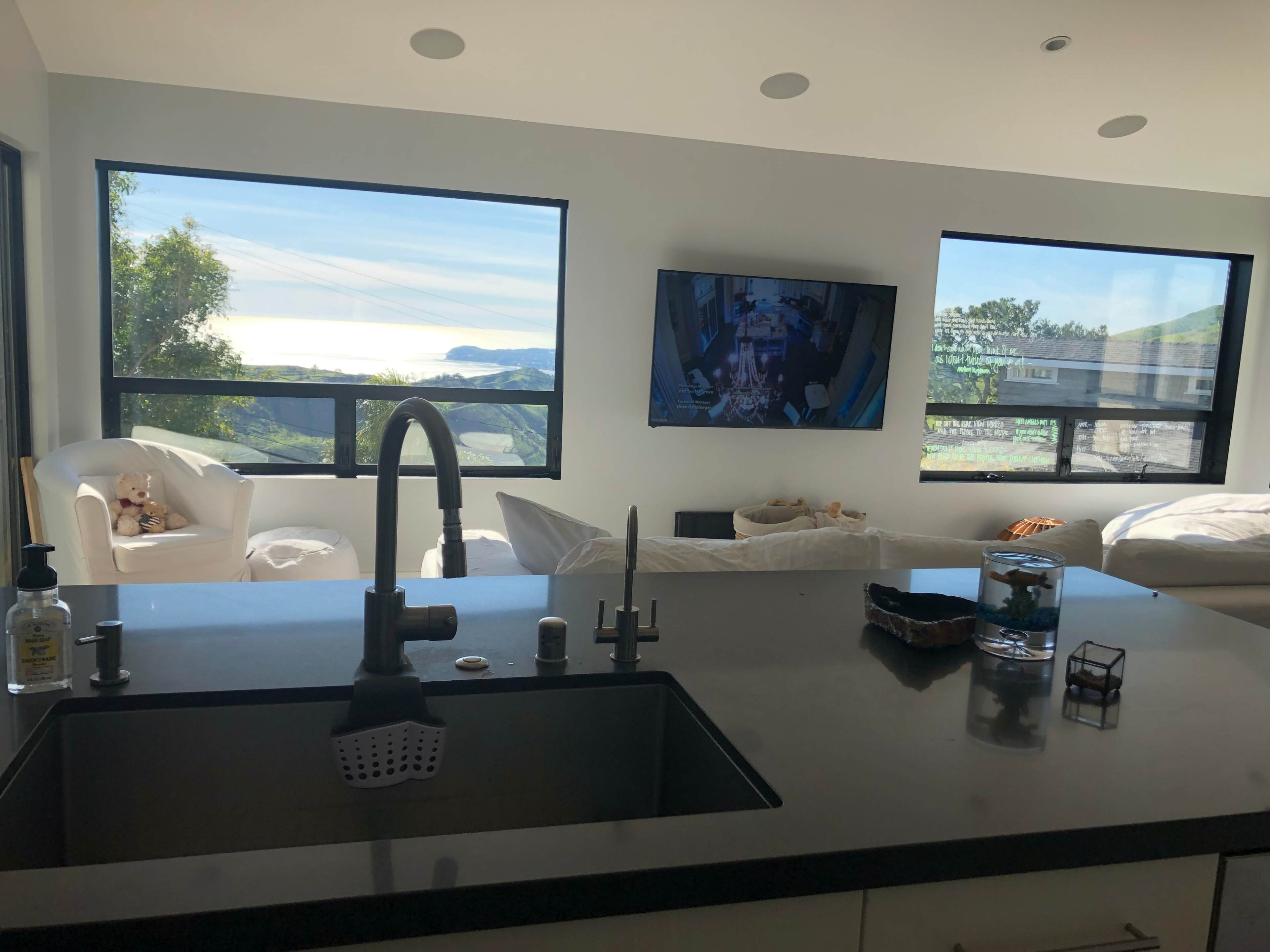 A modern kitchen view shows a sink in the foreground, with a television and seating area visible through large windows overlooking a scenic landscape.