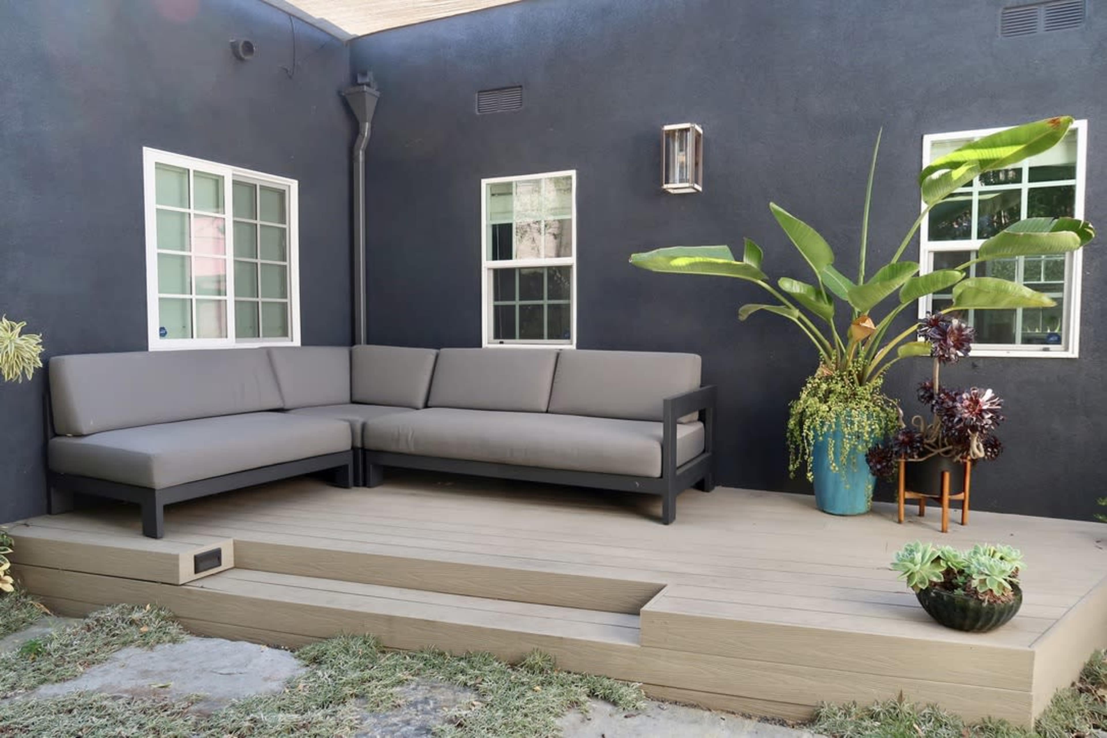 The image shows a modern outdoor seating area with a gray sectional sofa on a wooden platform, surrounded by potted plants and two windows.