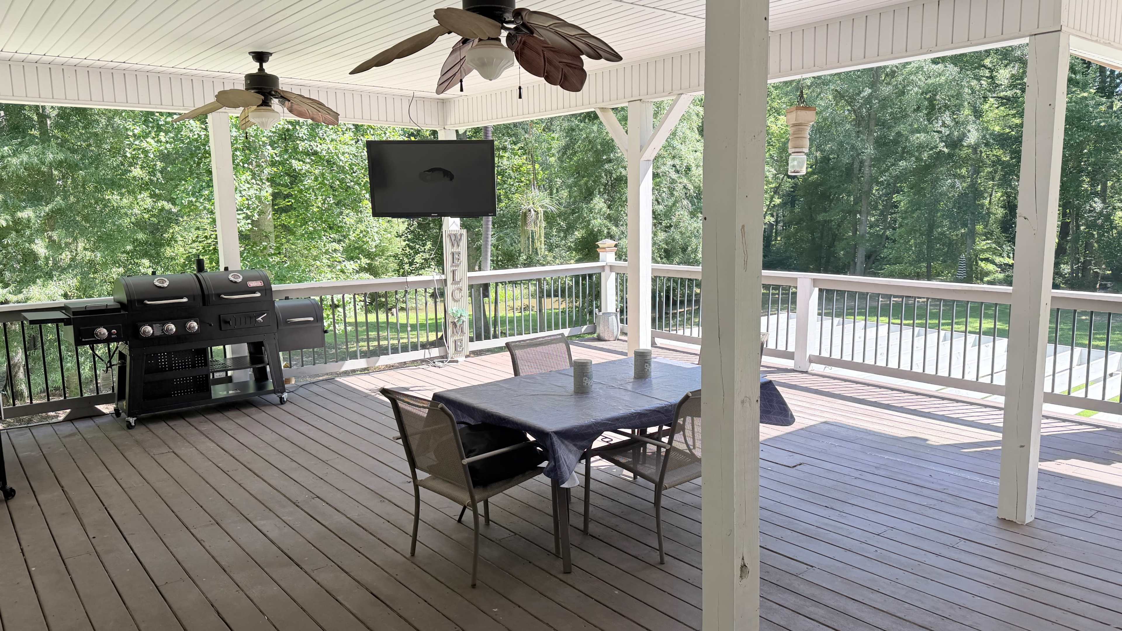 The image shows a covered outdoor deck with a dining table, a grill, and a television mounted on a post, surrounded by trees.