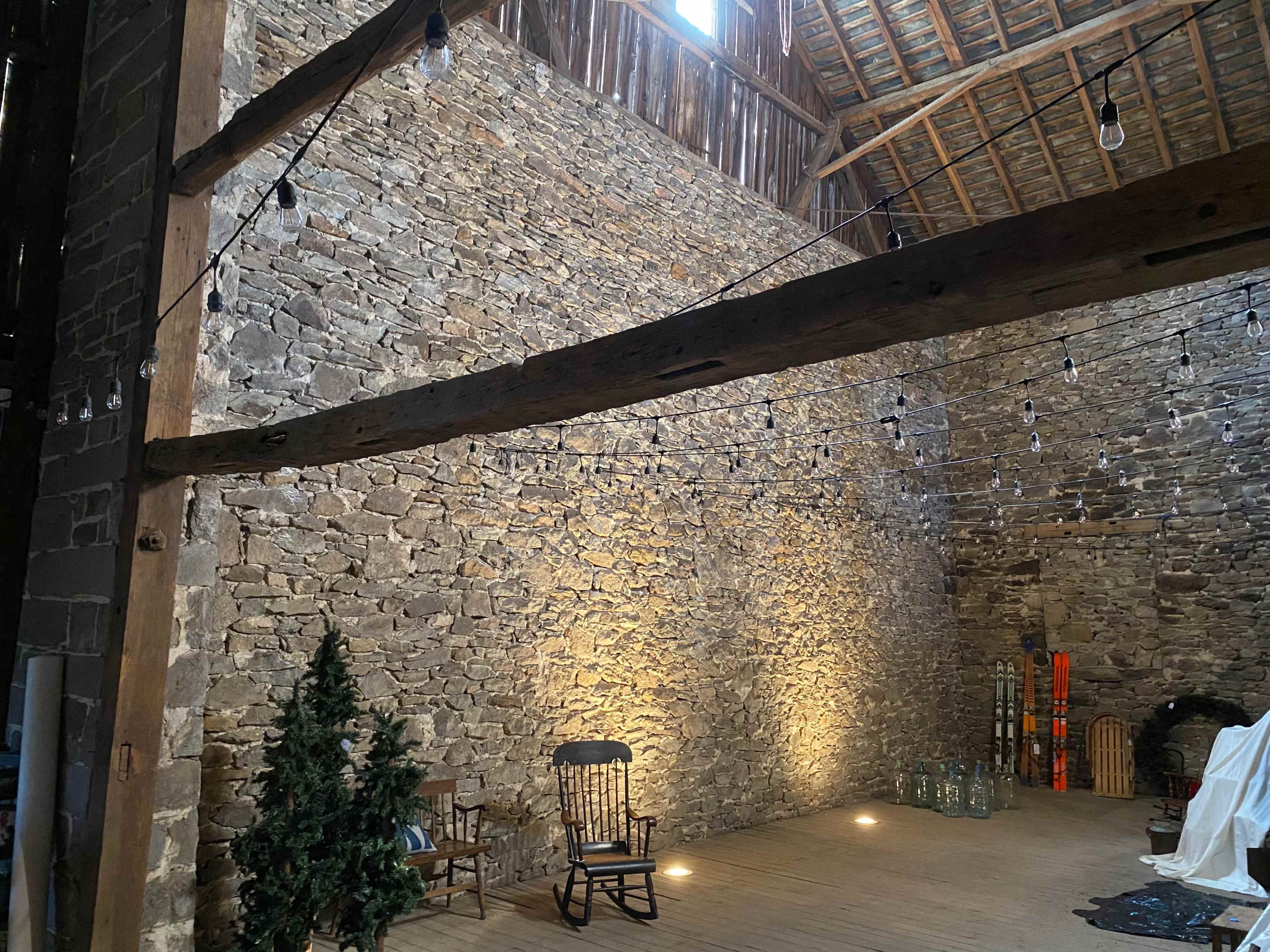 A rustic barn interior with a stone wall, wooden beams, and a wooden floor, decorated with string lights and a few pieces of furniture.
