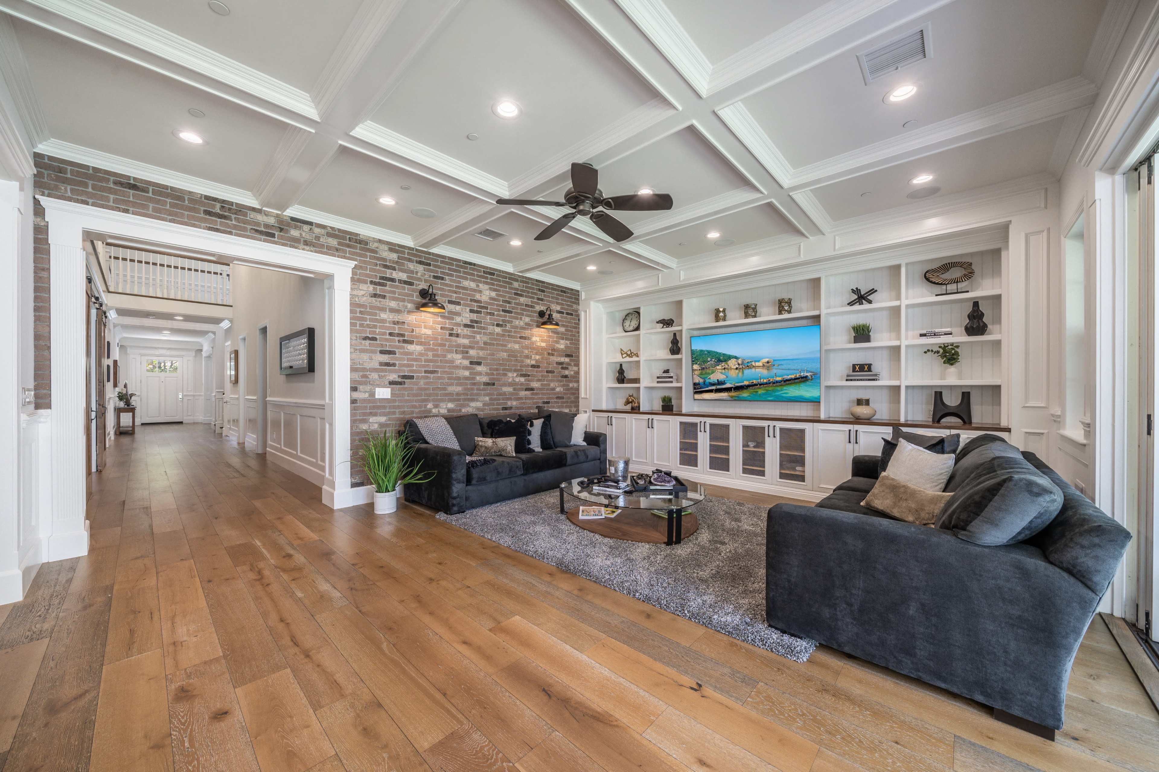 A spacious living room features a coffered ceiling, brick accent wall, and modern furnishings arranged around a central coffee table.
