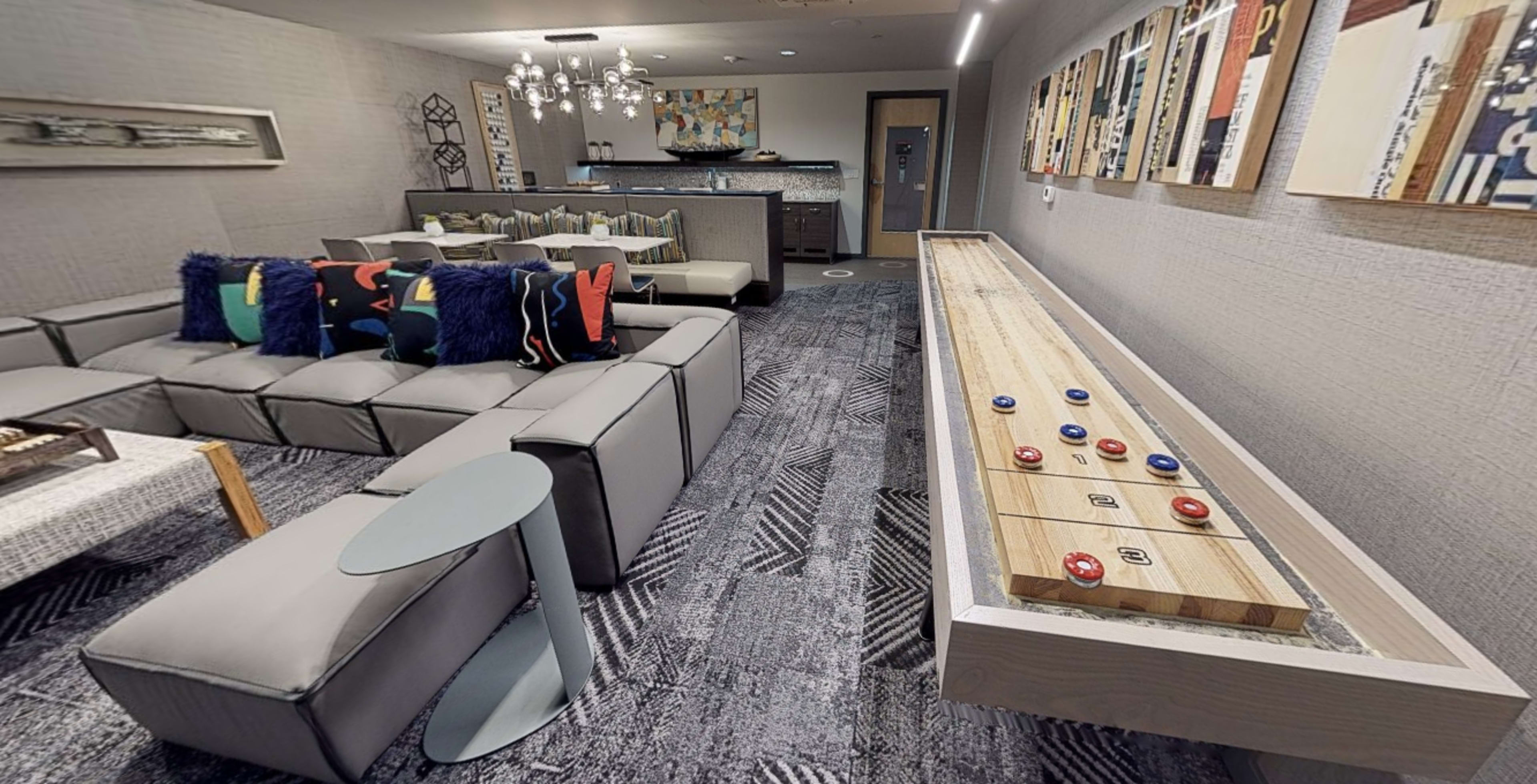 A modern lounge area featuring a large sectional sofa, colorful cushions, and a shuffleboard table along one wall.