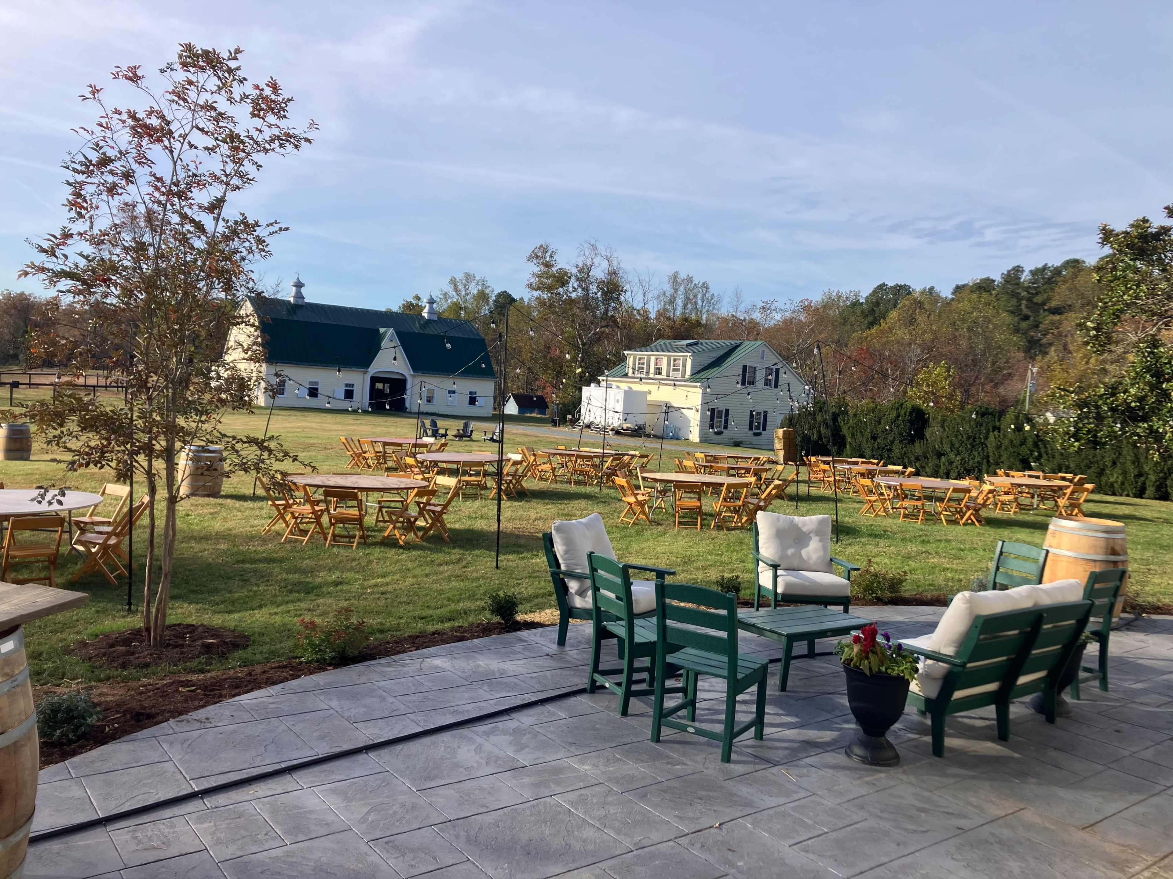 The image shows an outdoor event space with wooden tables and chairs arranged on a grassy area, surrounded by trees and buildings.