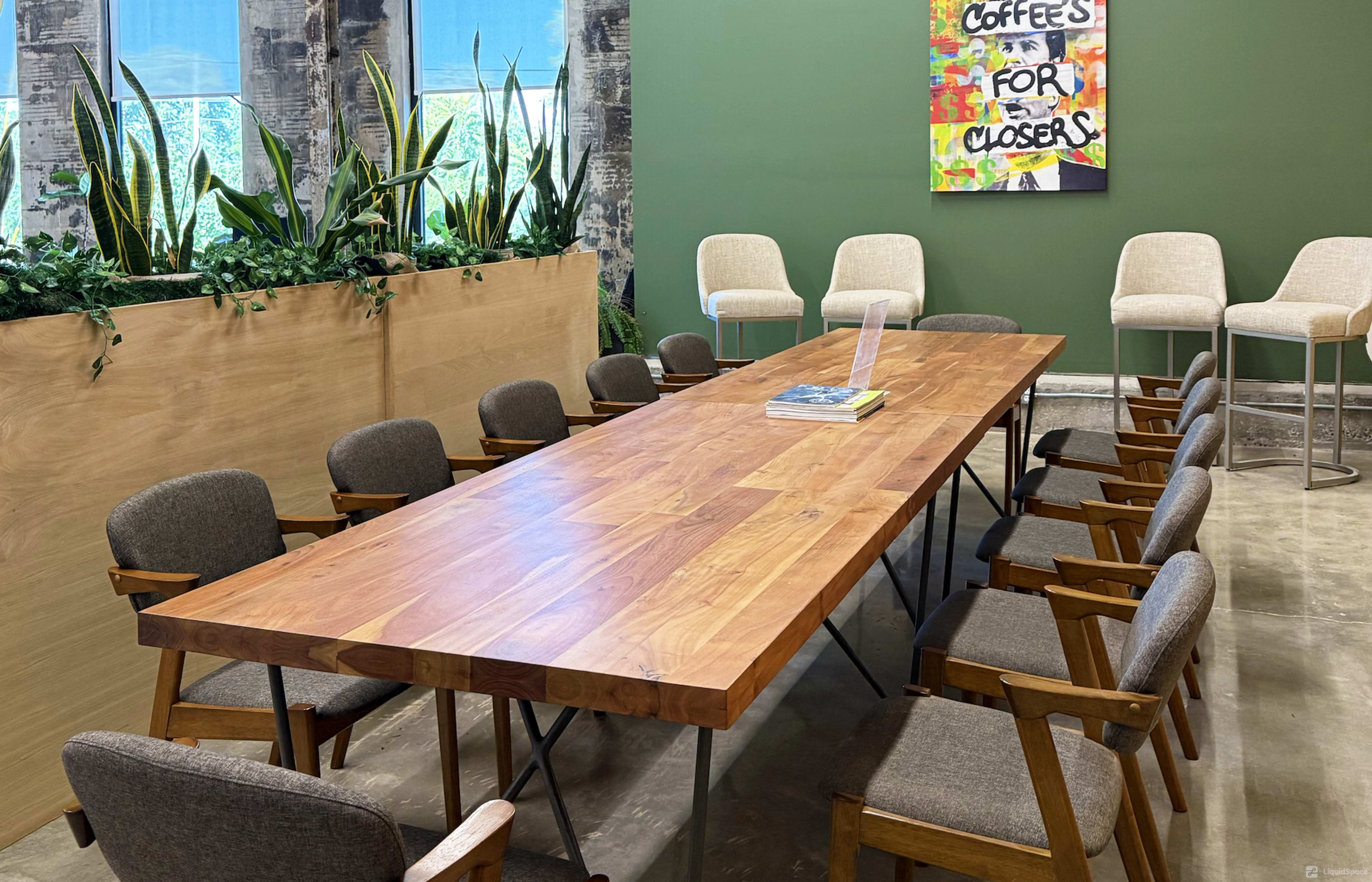 A long wooden table with wooden and upholstered chairs is set in a room featuring green walls and large windows, alongside a decorative plant display and a framed sign that reads "Coffee for Closers."