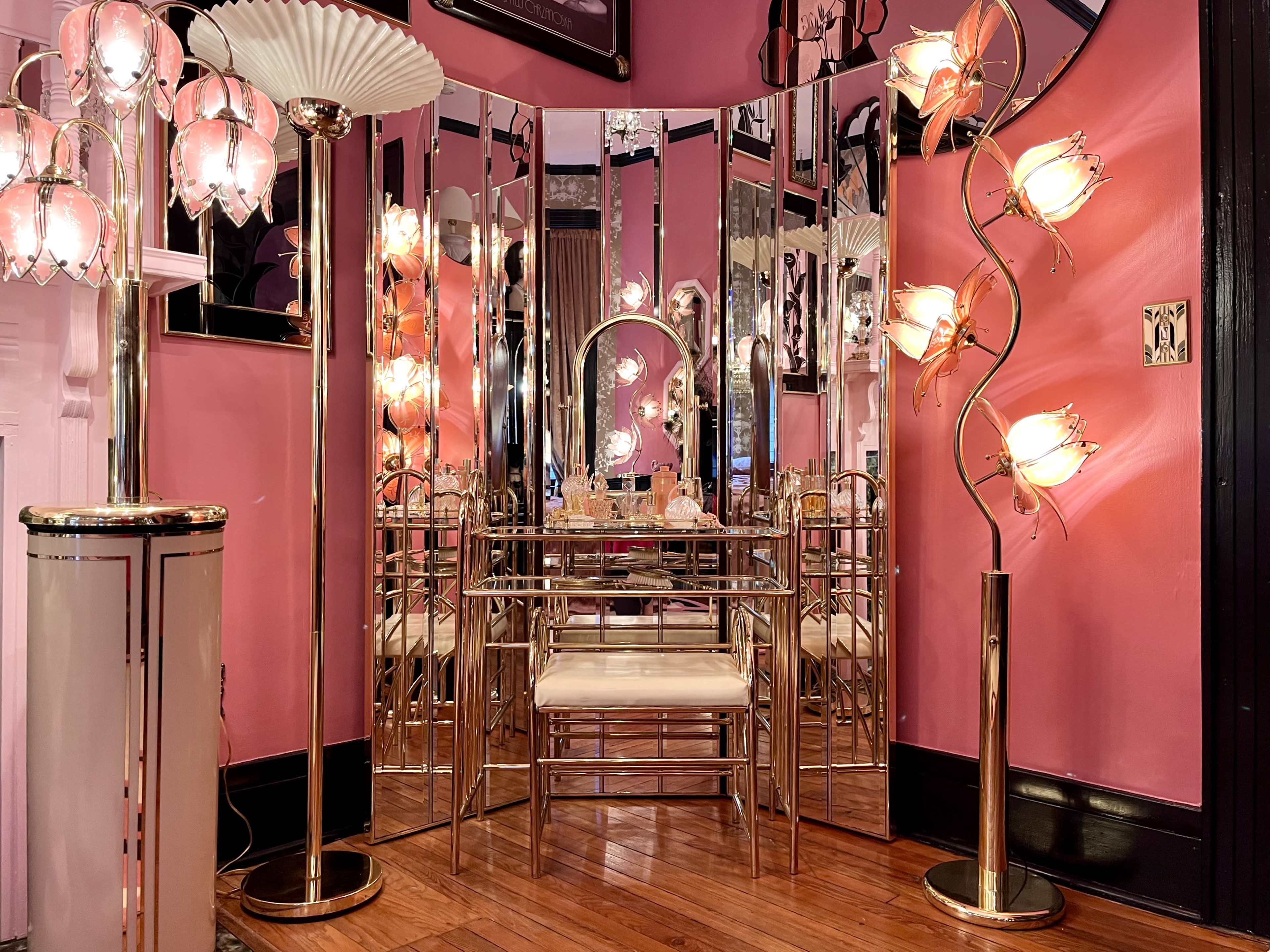 The image shows a room with pink walls featuring a mirrored dressing area, a circular mirror, and decorative lighting, including a standing lamp and a spiral lamp with floral designs.