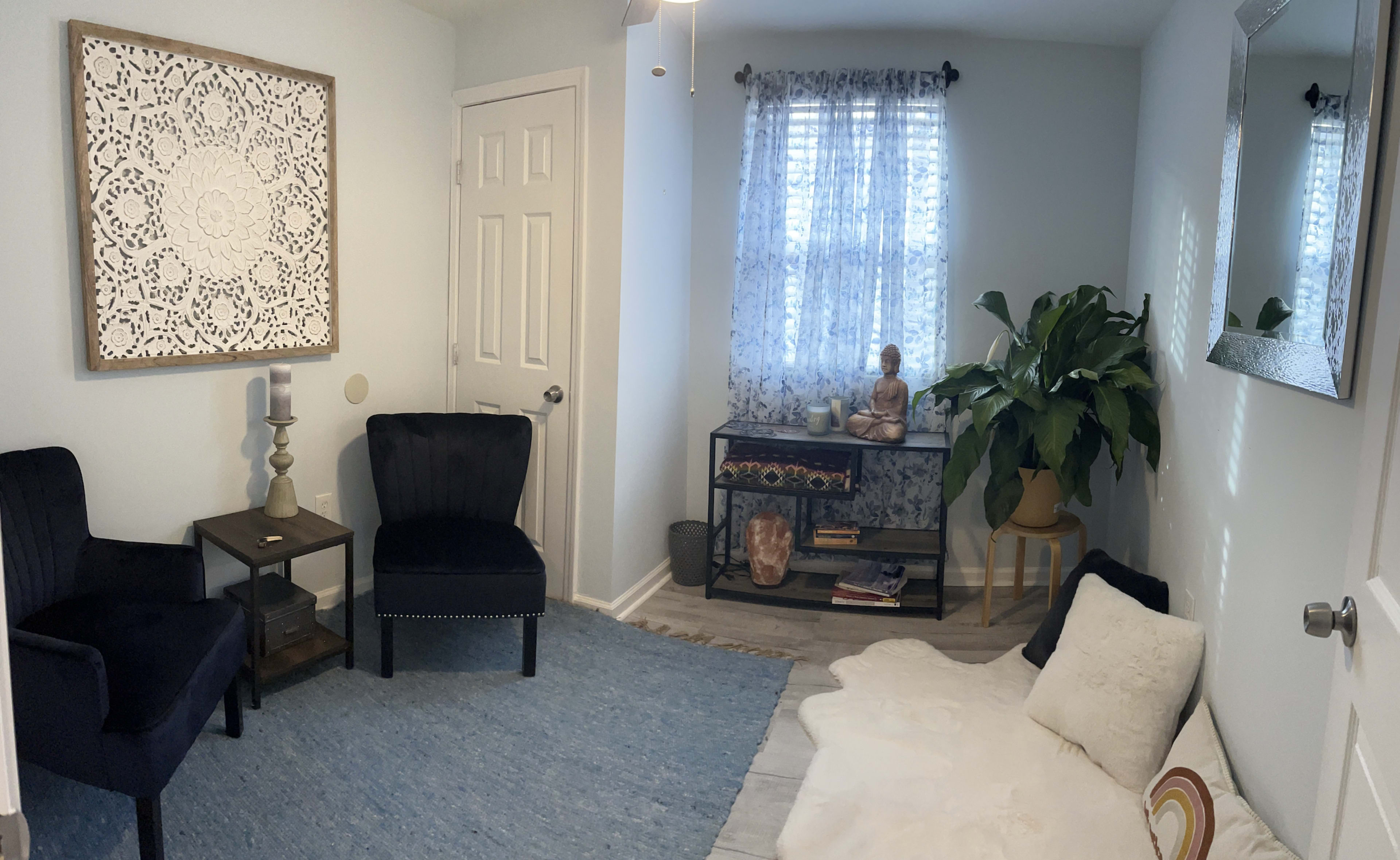 A small, well-lit room features two black chairs, a white area rug, and a bookcase with decorative items, including a statue of a Buddha and a large plant near a window.