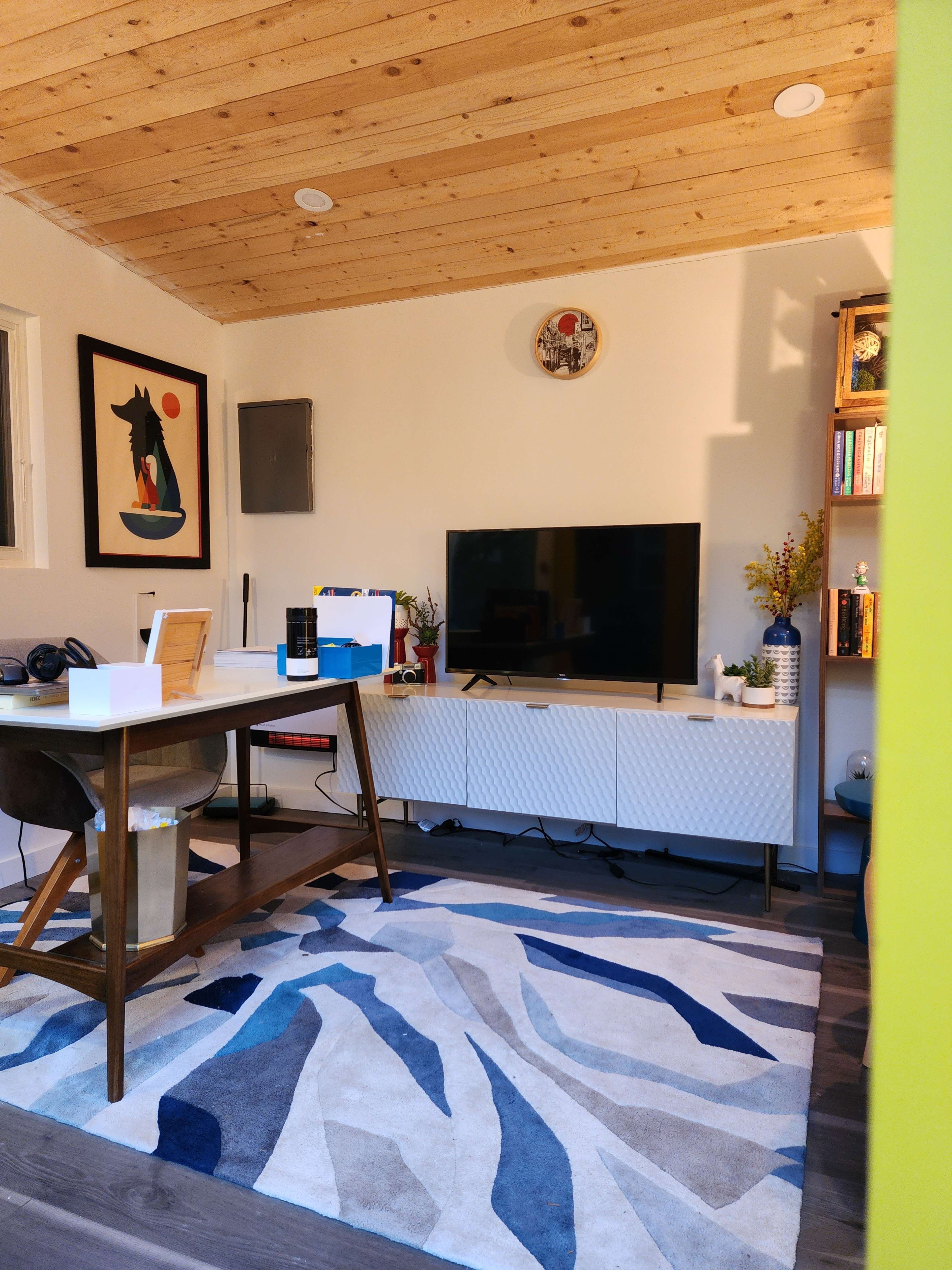 A cozy room features a wooden ceiling, a television on a white cabinet, a desk with a computer, and decorative items on shelves.