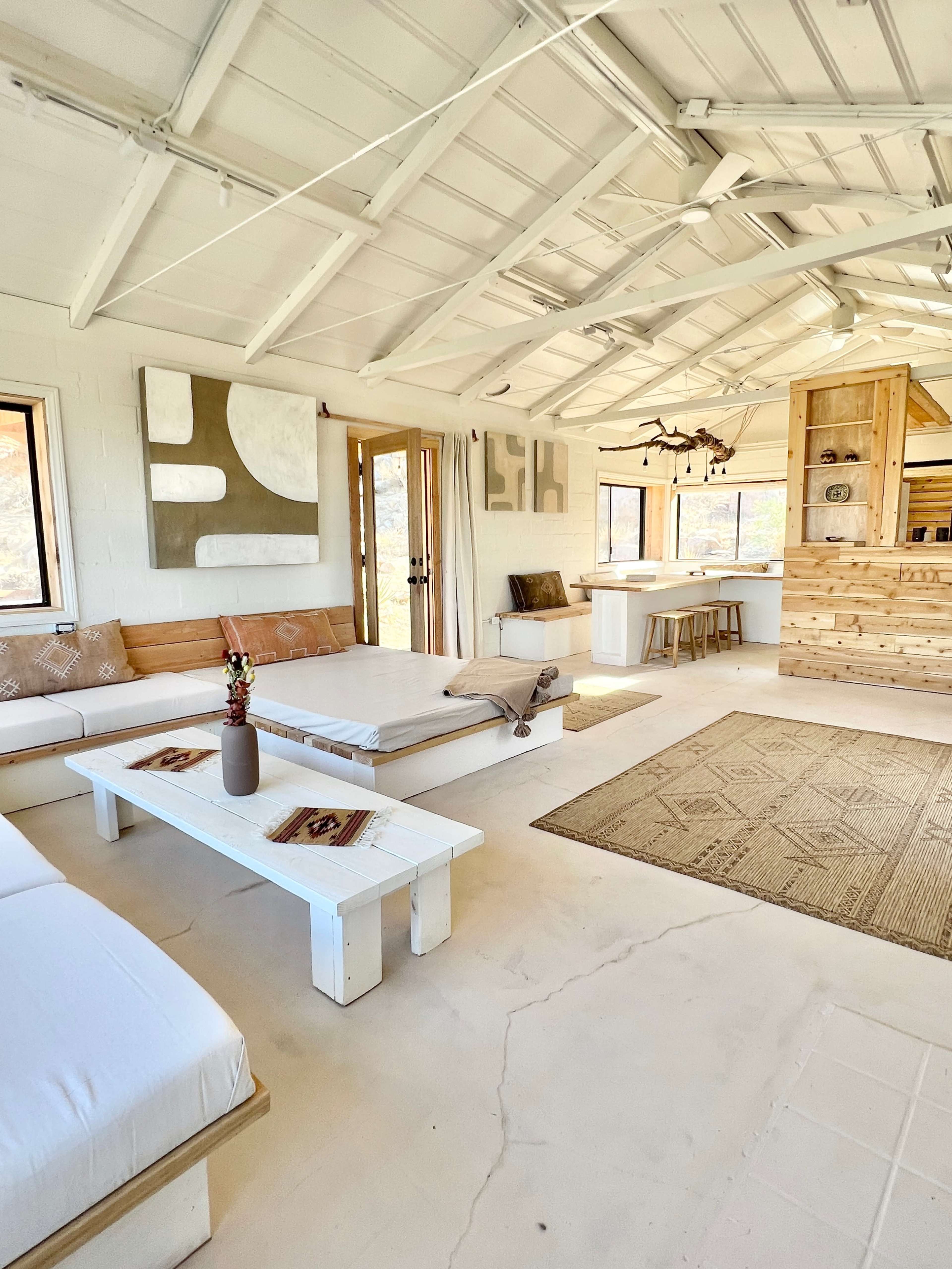 A spacious, well-lit interior of a minimalist living area with a few beige and white furnishings, a wooden kitchen area, and a large rug on the floor.
