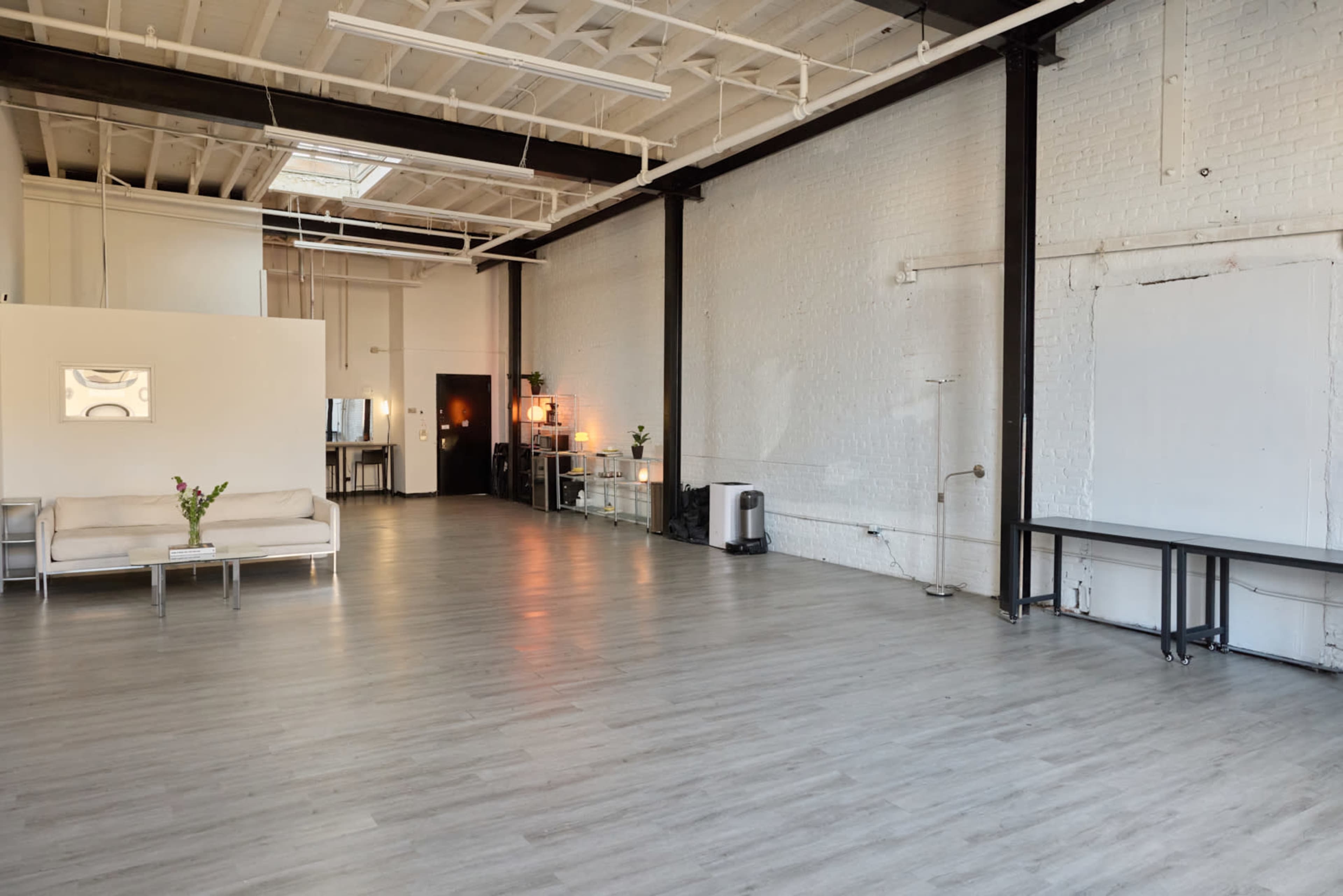 The image shows a spacious, minimalist interior with light wooden flooring and white brick walls, featuring a couch, a small table, and a few decorative items.