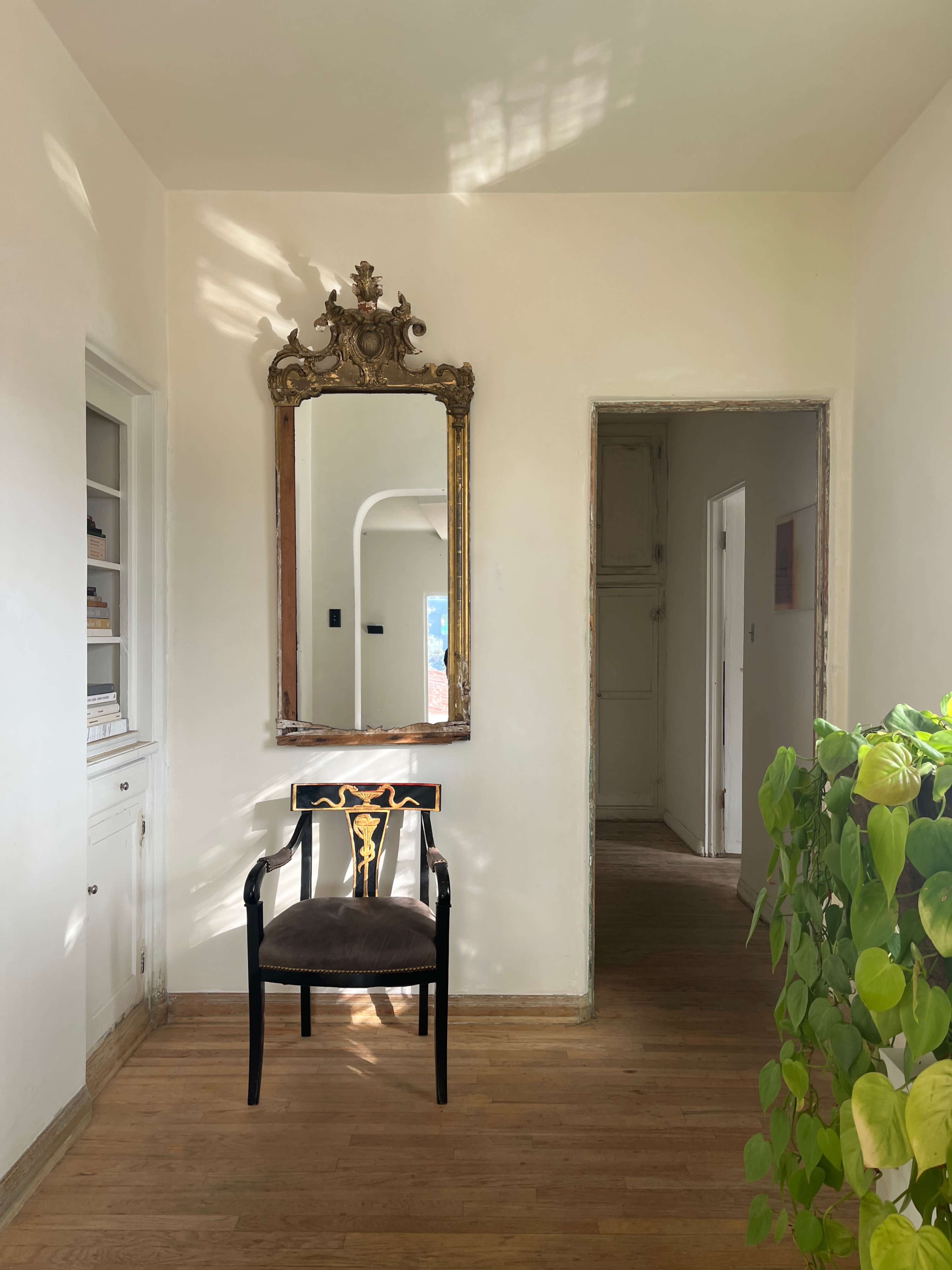 A bright hallway with a chair, a decorative mirror on the wall, and a leafy plant in the corner.