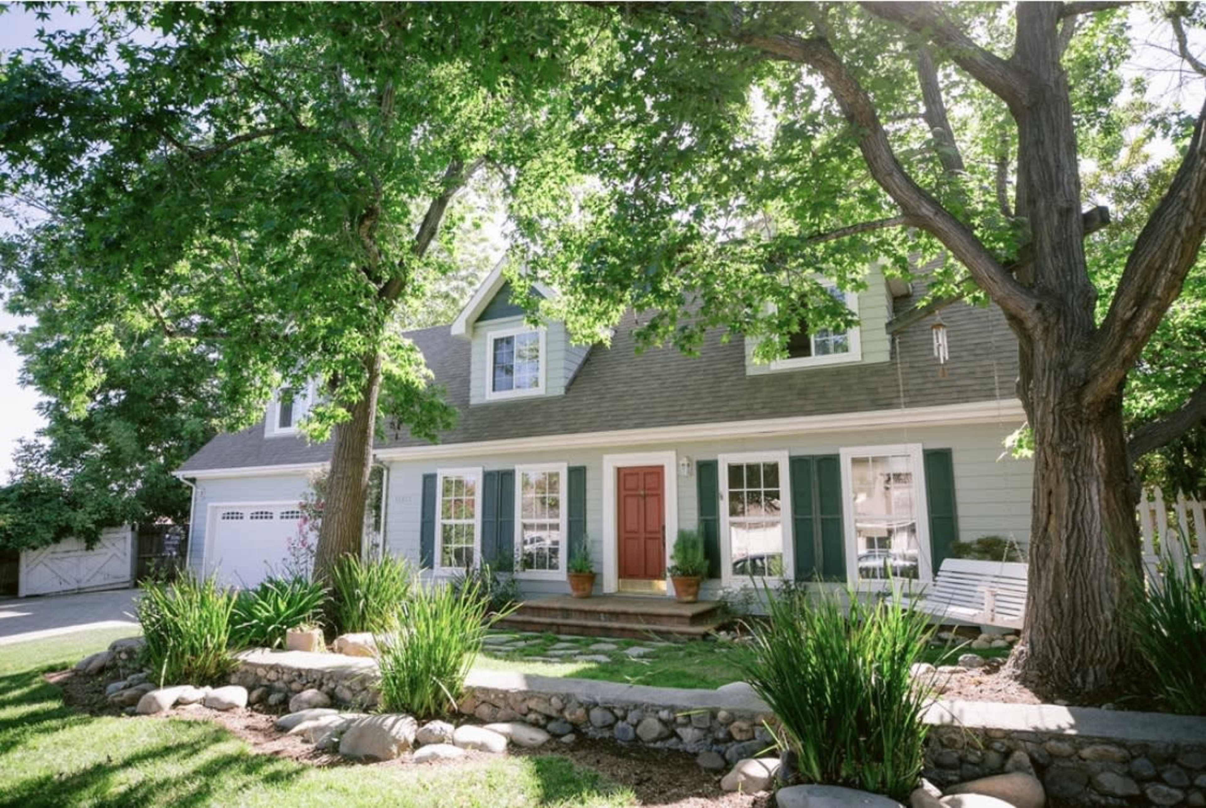 A two-story house with green shutters and a red front door, surrounded by trees, a landscaped garden, and a stone pathway.